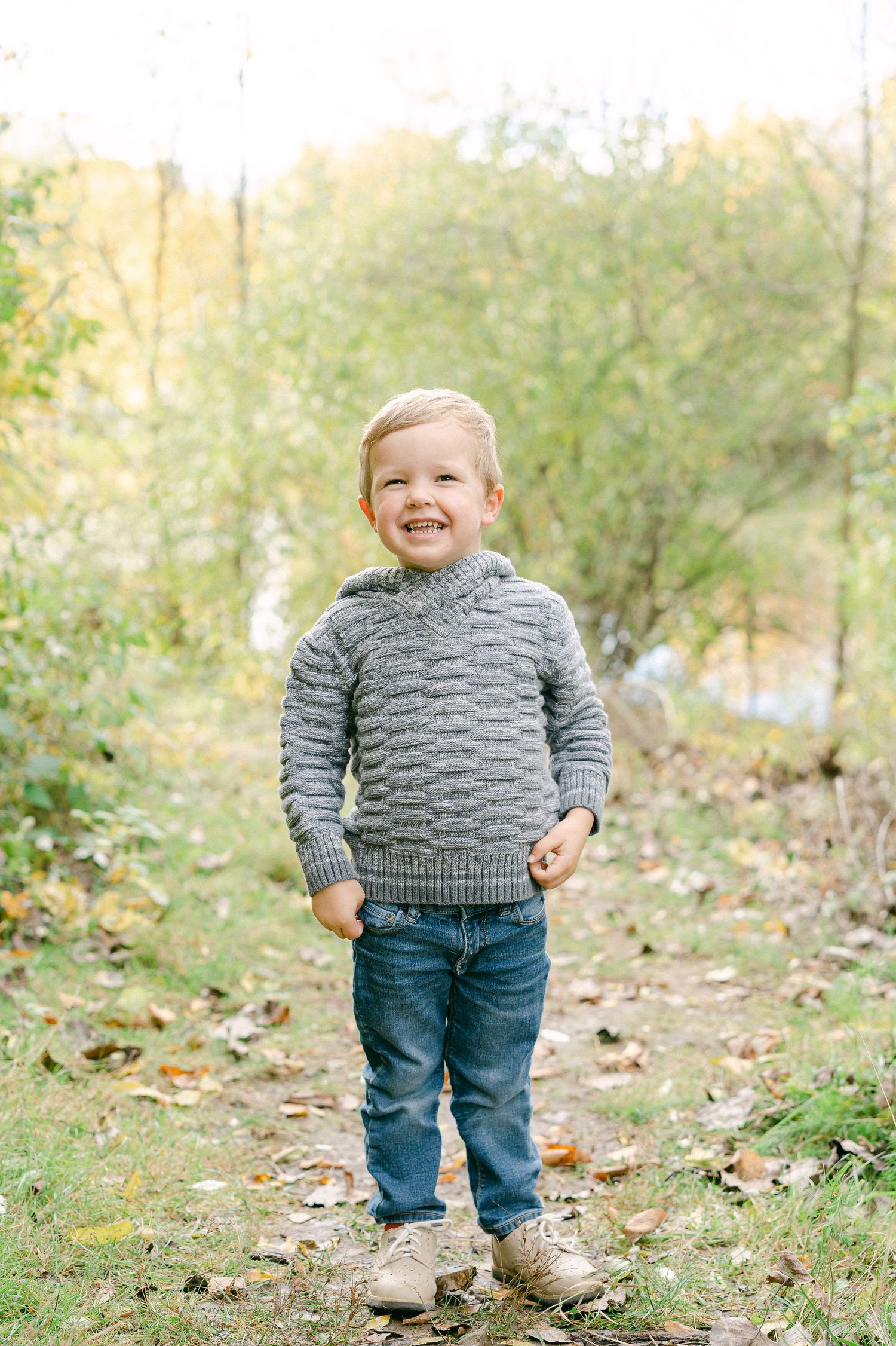 A small figure in a gray striped sweater and jeans stands among fallen leaves on a grassy path.