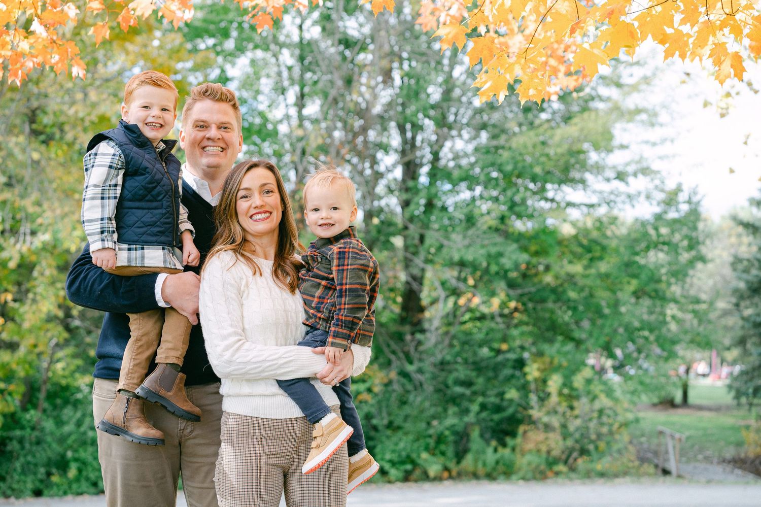 A family poses together outdoors in autumn with golden leaves overhead and green trees in the background.