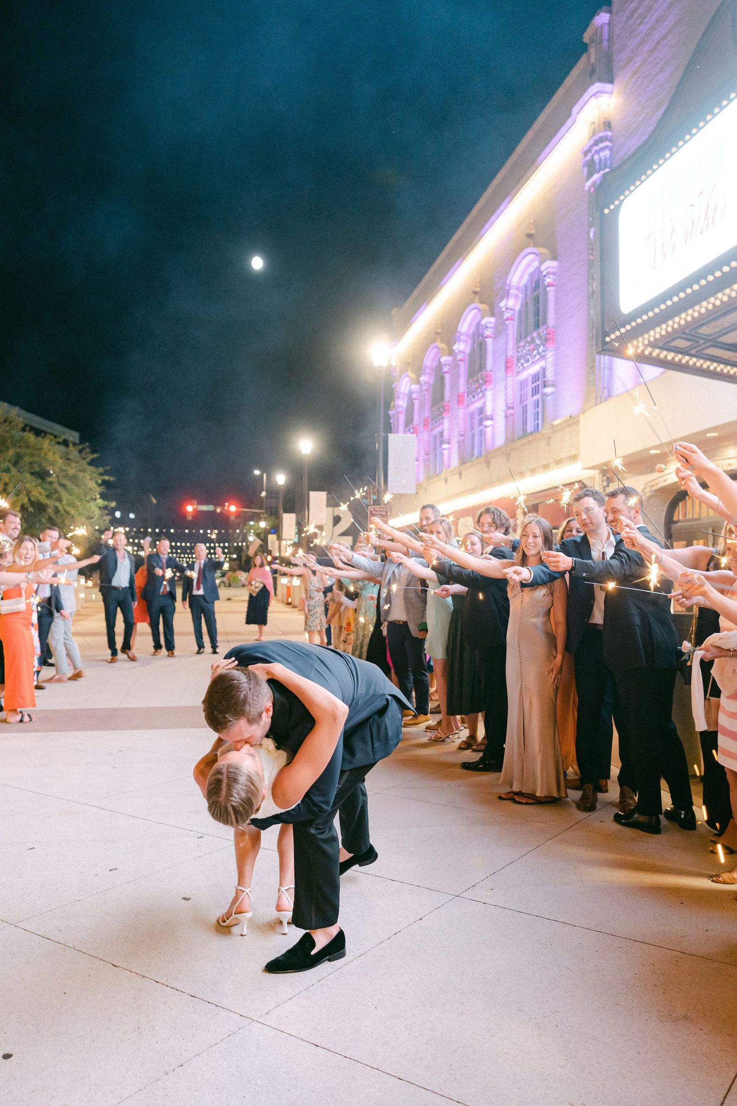 Couple shares romantic dip kiss during sparkler send-off outside illuminated venue at night wedding celebration in South Bend, IN