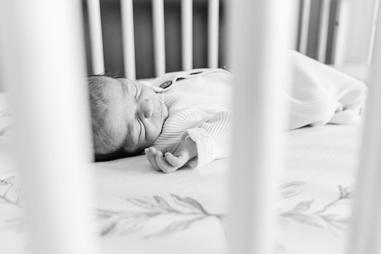 A black and white close-up photo shows a baby sleeping peacefully between white crib slats.