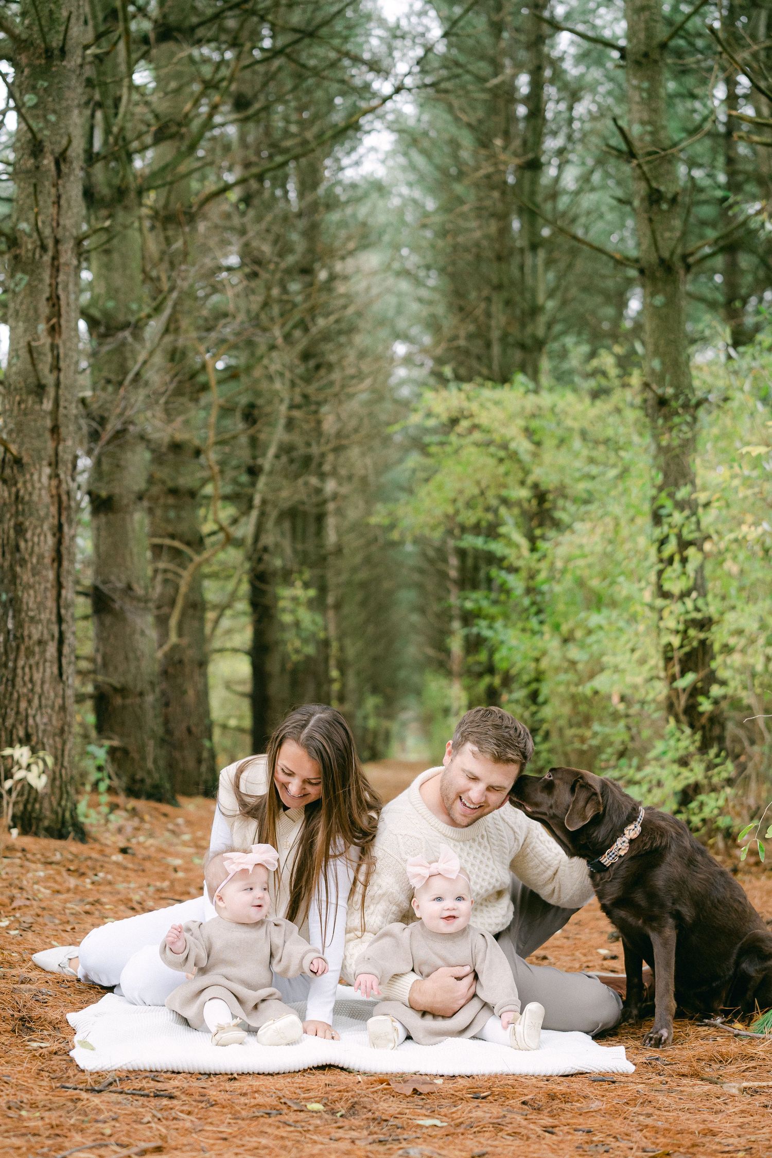 Family lying on white blanket in pine forest with their black dog enjoying outdoor photo session.
