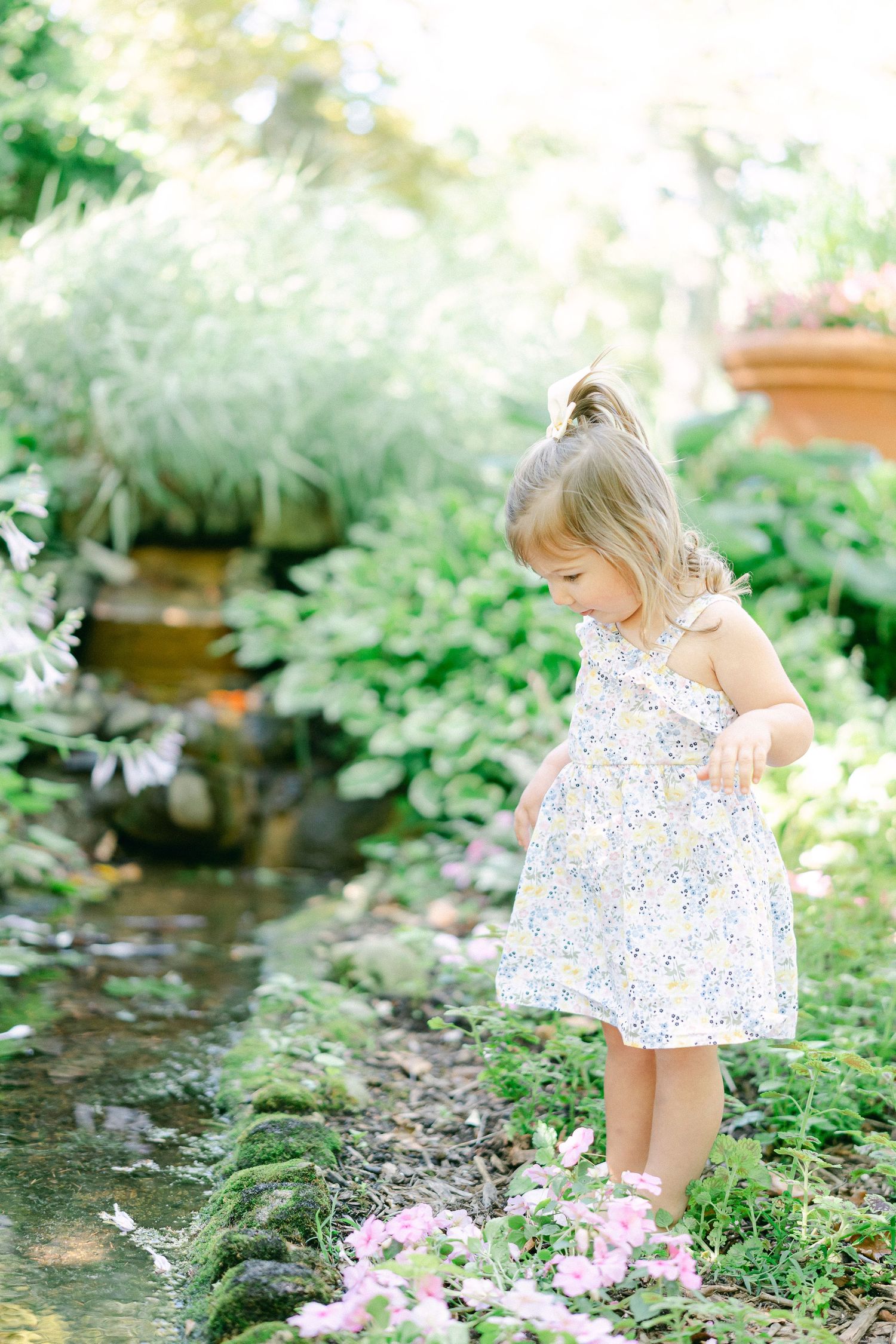 A small child in a floral dress explores near a garden stream surrounded by lush greenery and pink flowers.