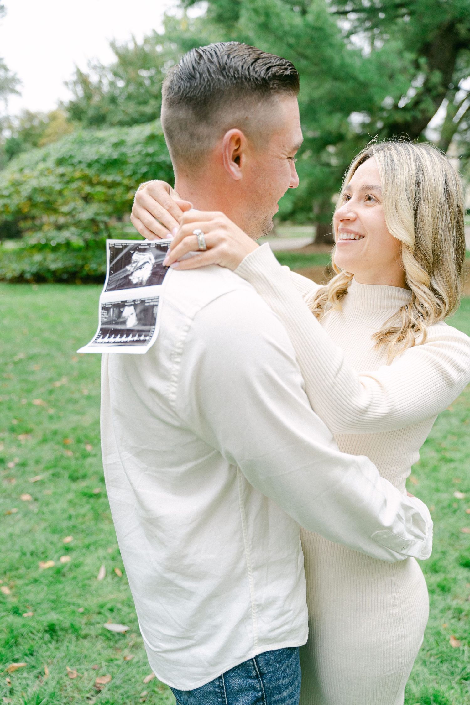 A couple in white outfits dances together in a park setting while holding a small photograph.