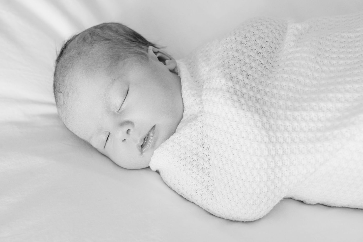 Peaceful sleeping newborn baby wrapped in white textured blanket on light bedding.