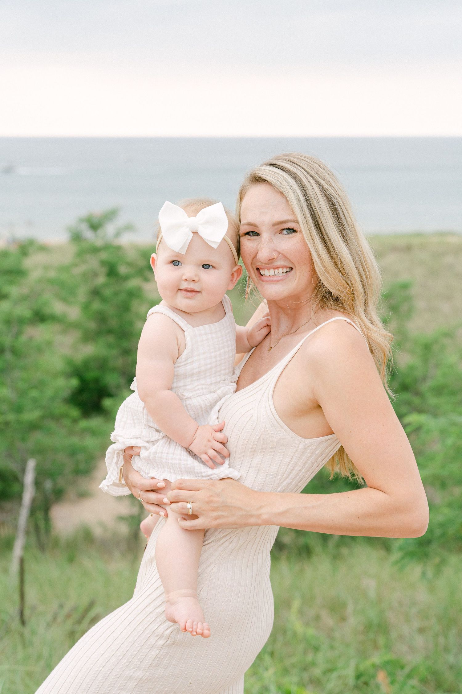 Mother and baby in matching white outfits share tender moments against a scenic coastal backdrop.