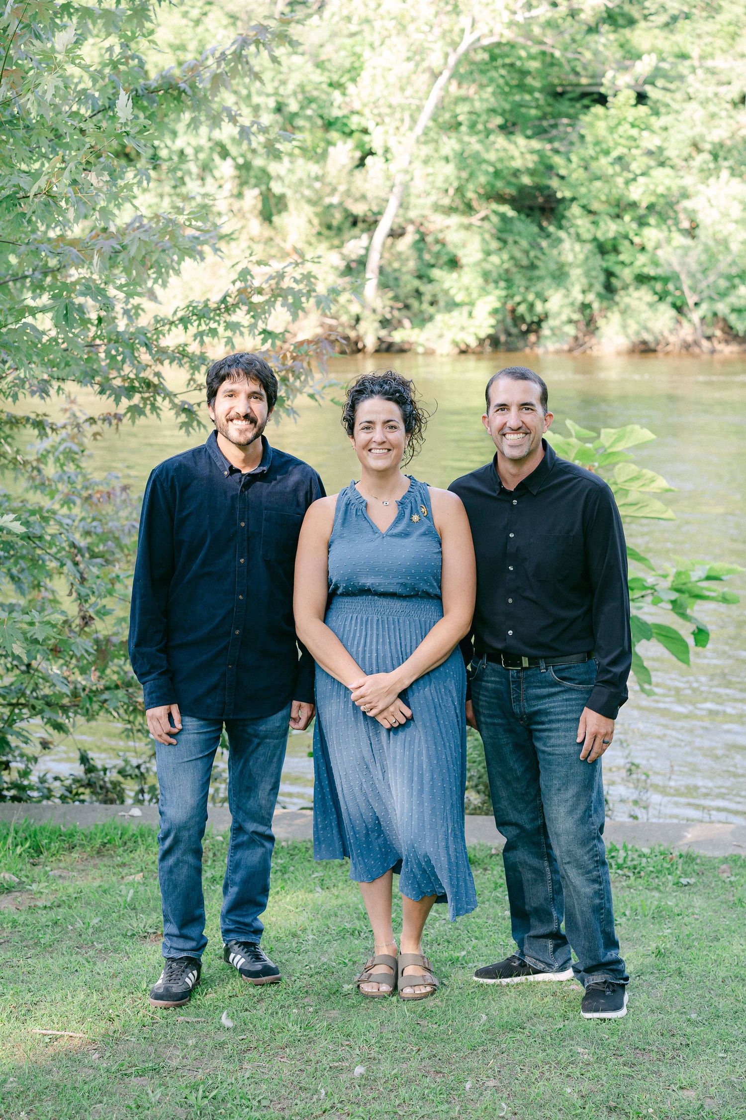 Three people stand together on grass near a river, with the center person wearing a blue dress.