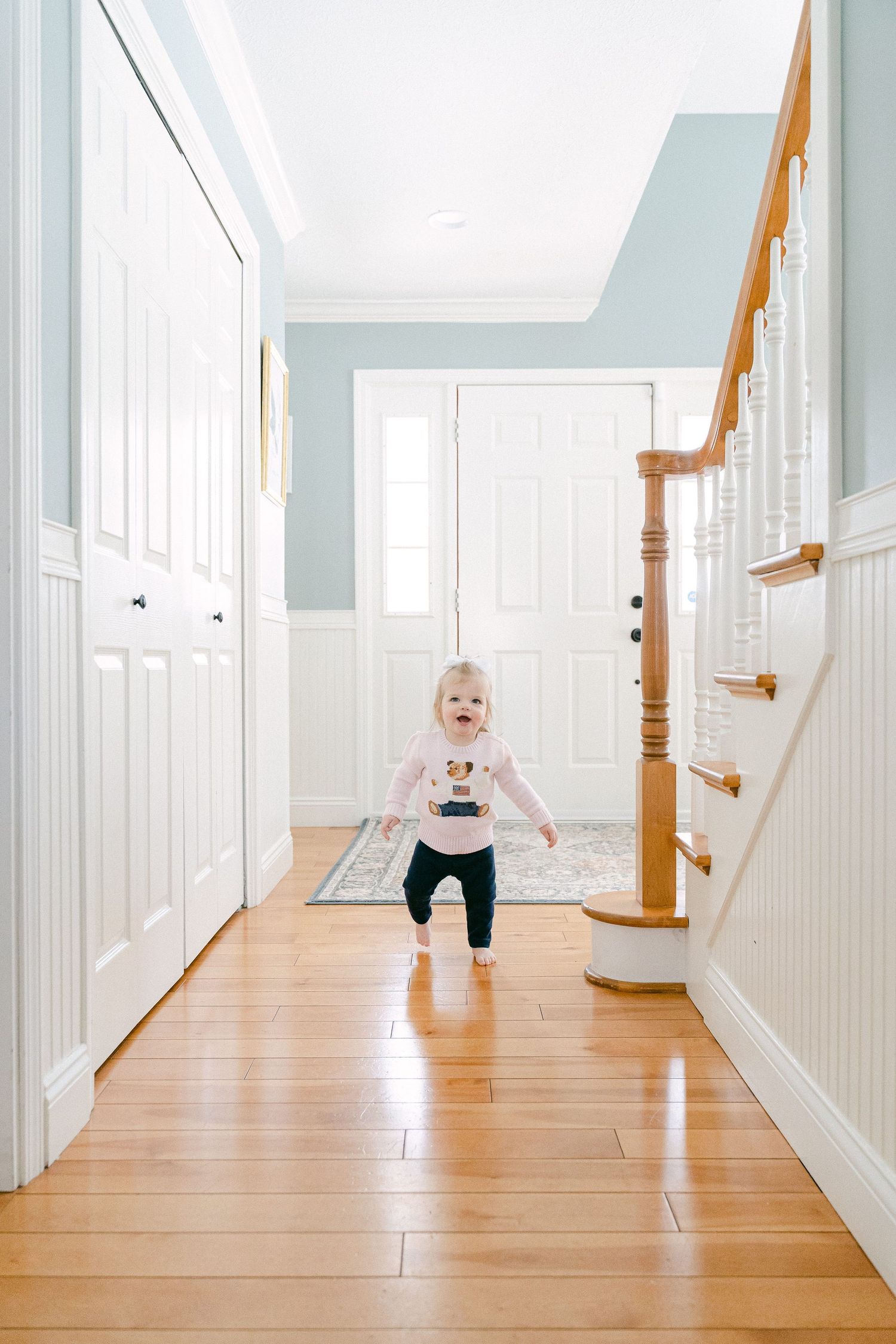 Young child running and playing in a bright hallway with white walls and hardwood flooring.