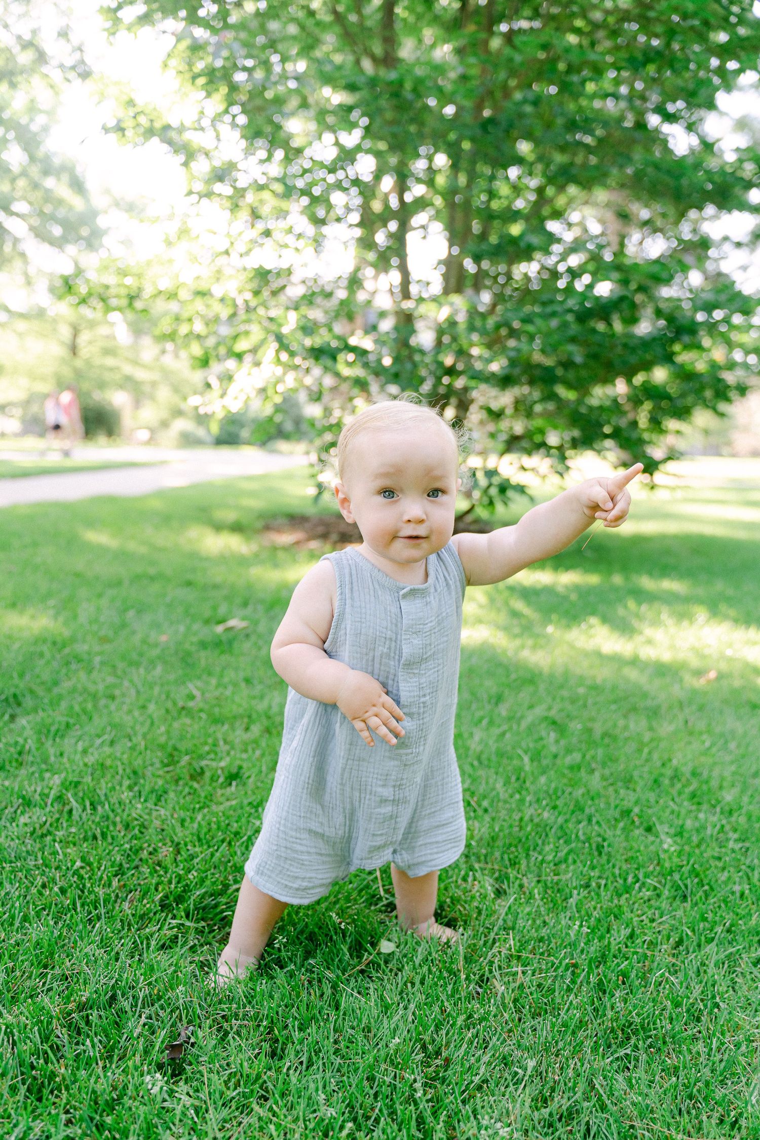 A baby in a grey romper takes wobbly steps on lush green grass during a sunny summer day.