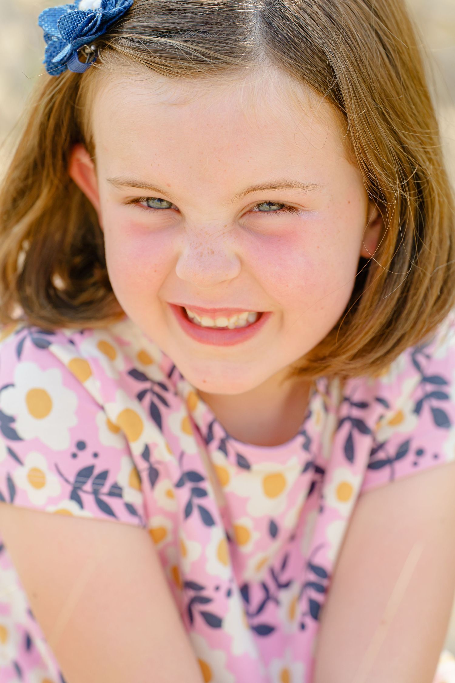 A cheerful child wearing a pink floral dress and blue hair accessory smiles brightly at the camera.