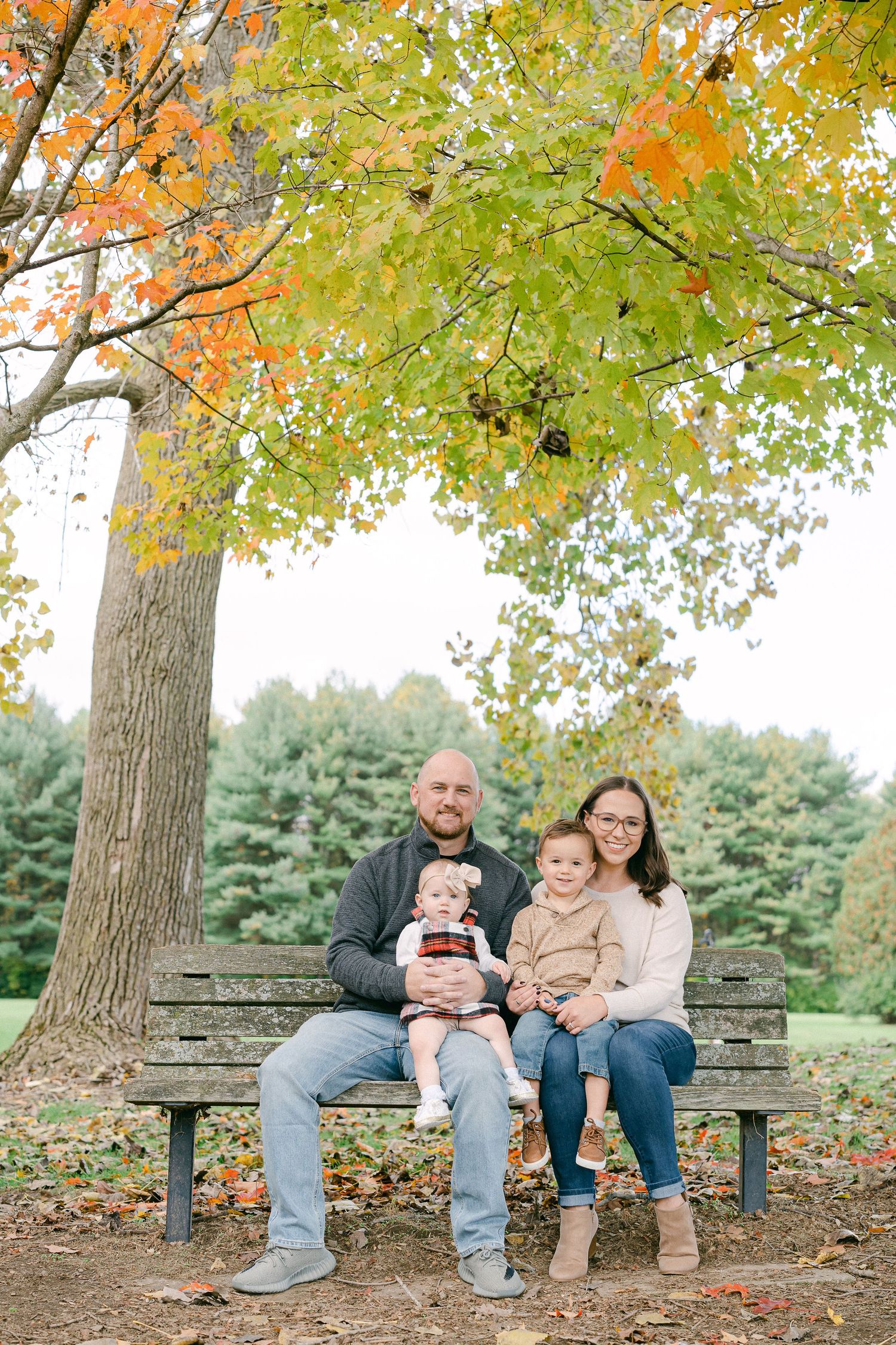 A family sits together on a stone bench under colorful autumn trees in a park setting.