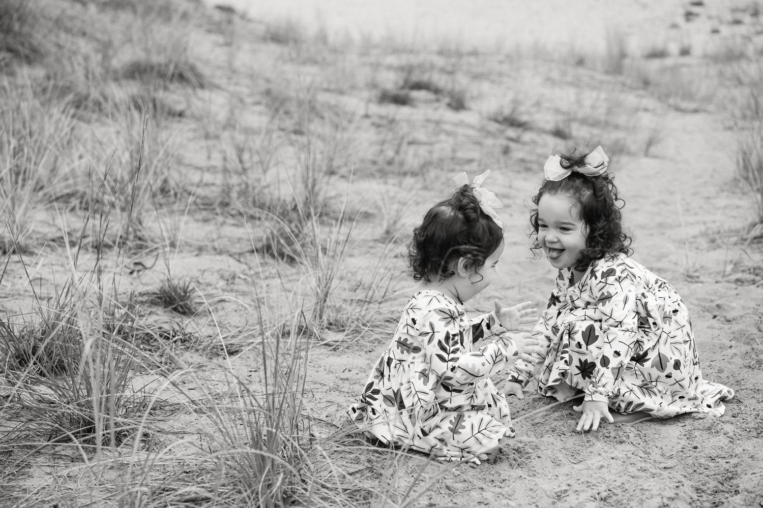 Two children in matching floral dresses sit together on sandy ground in a black and white photograph.