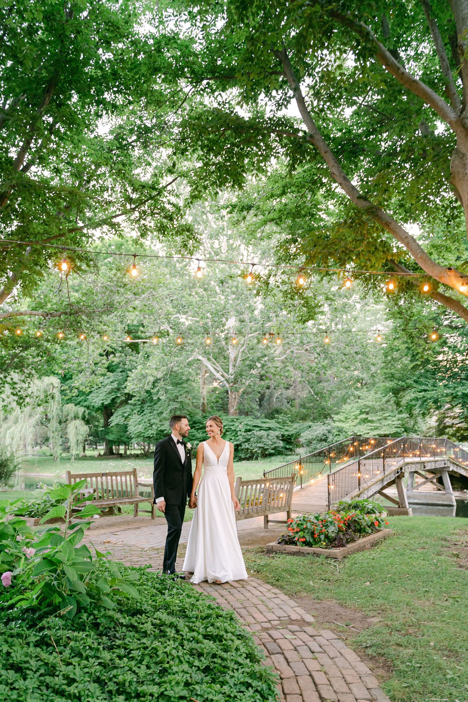 Bride and groom walk a brick path in a lush garden with string lights and a wooden bridge backdrop in South Bend,  IN