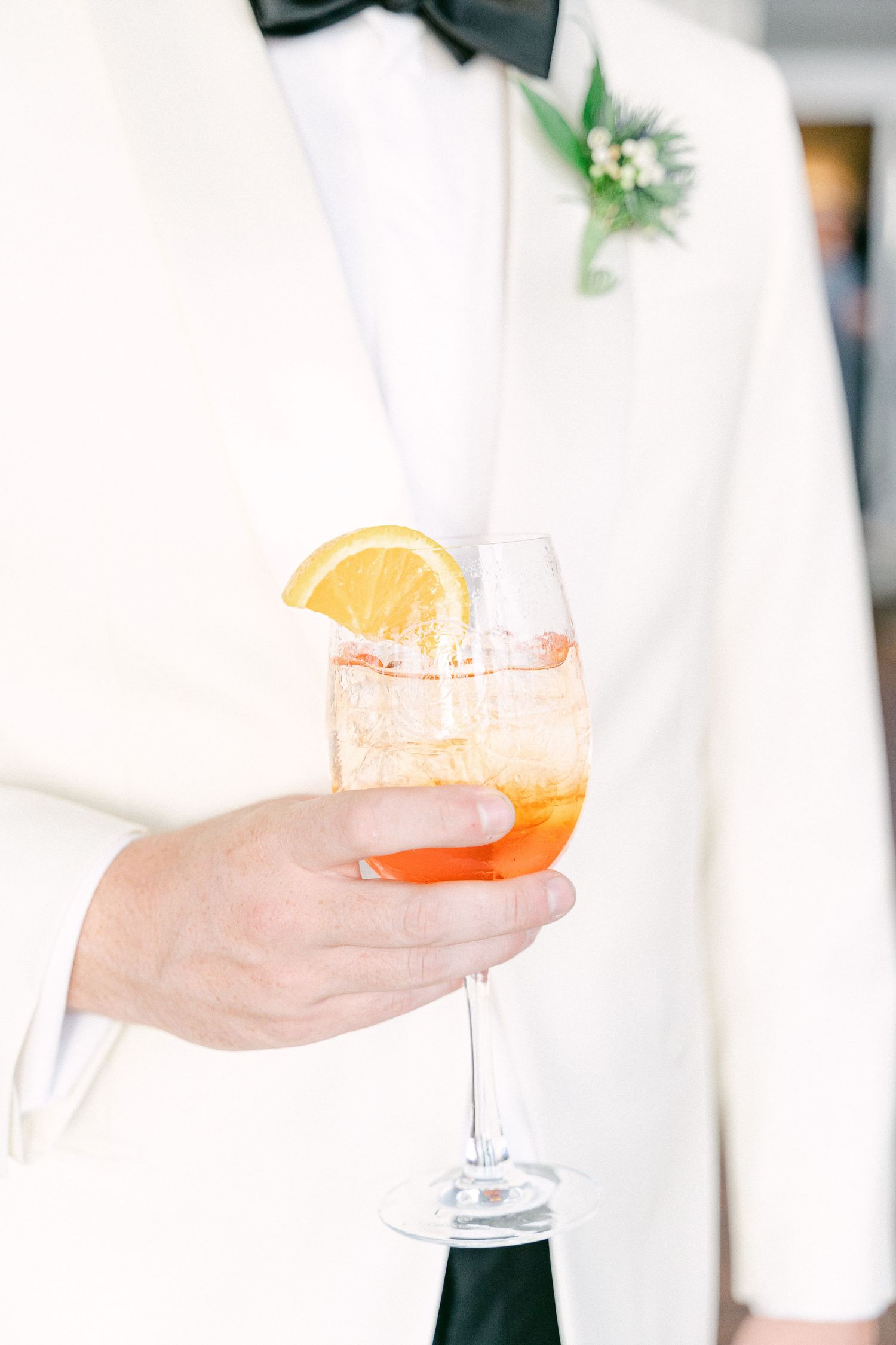 Groom in white tuxedo holding an Aperol Spritz cocktail garnished with an orange slice at a wedding in Cape Cod