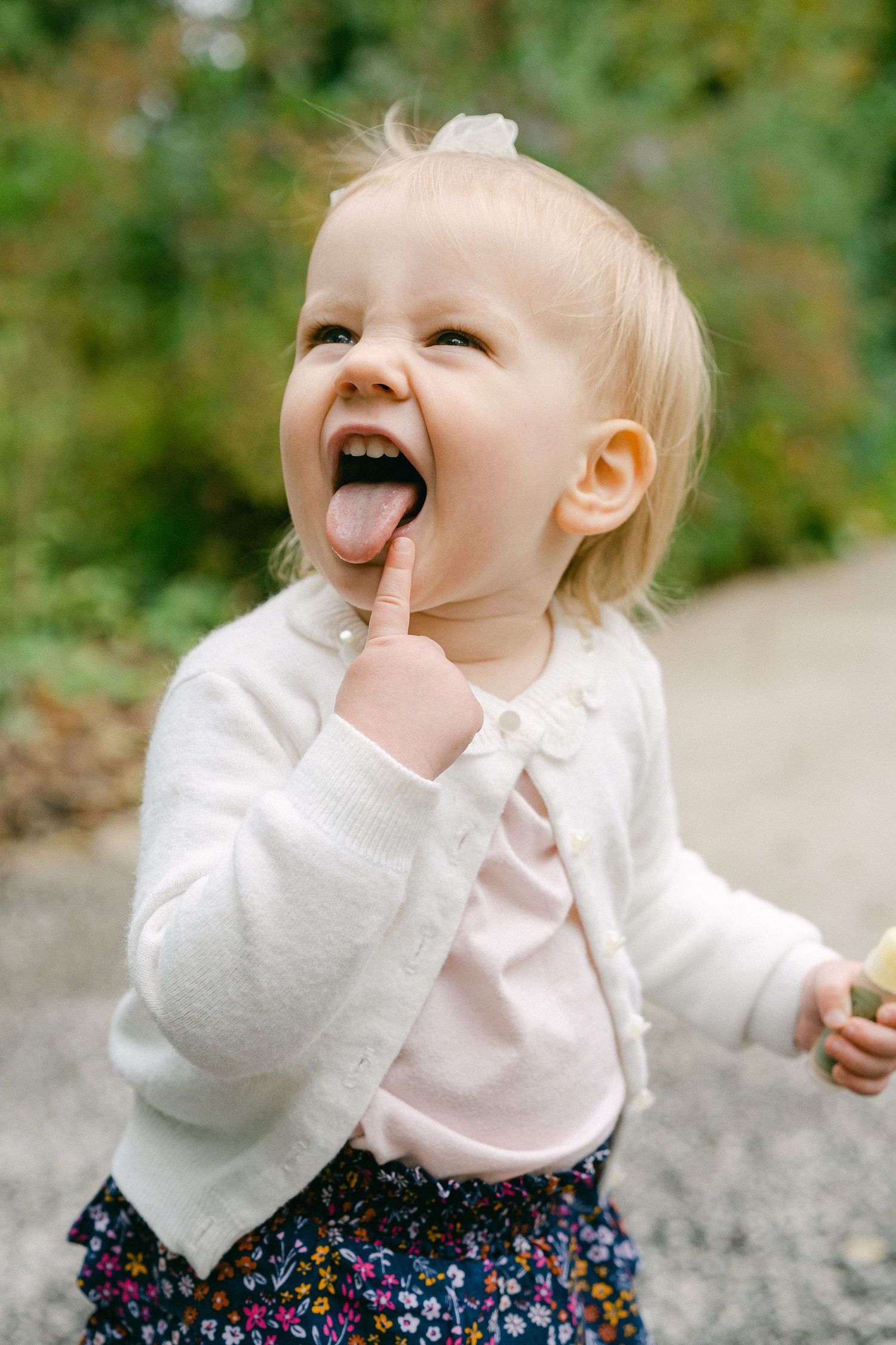 Child in white sweater and floral skirt making silly expressions while sitting outdoors.