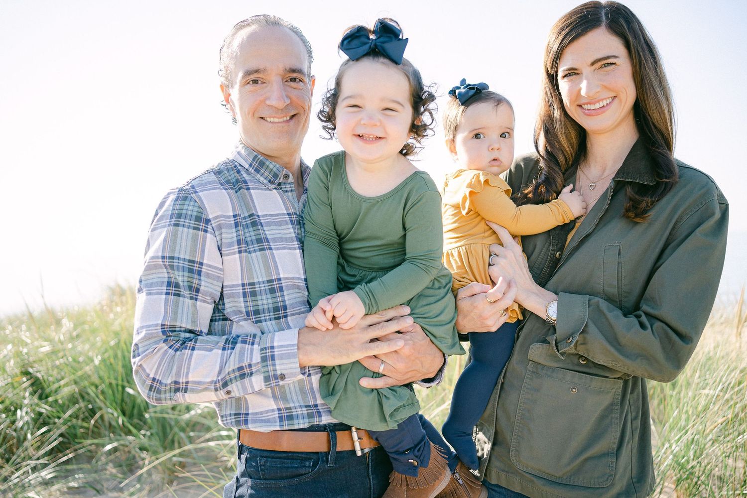 A happy family of four stands together outdoors in a grassy field on a sunny day.