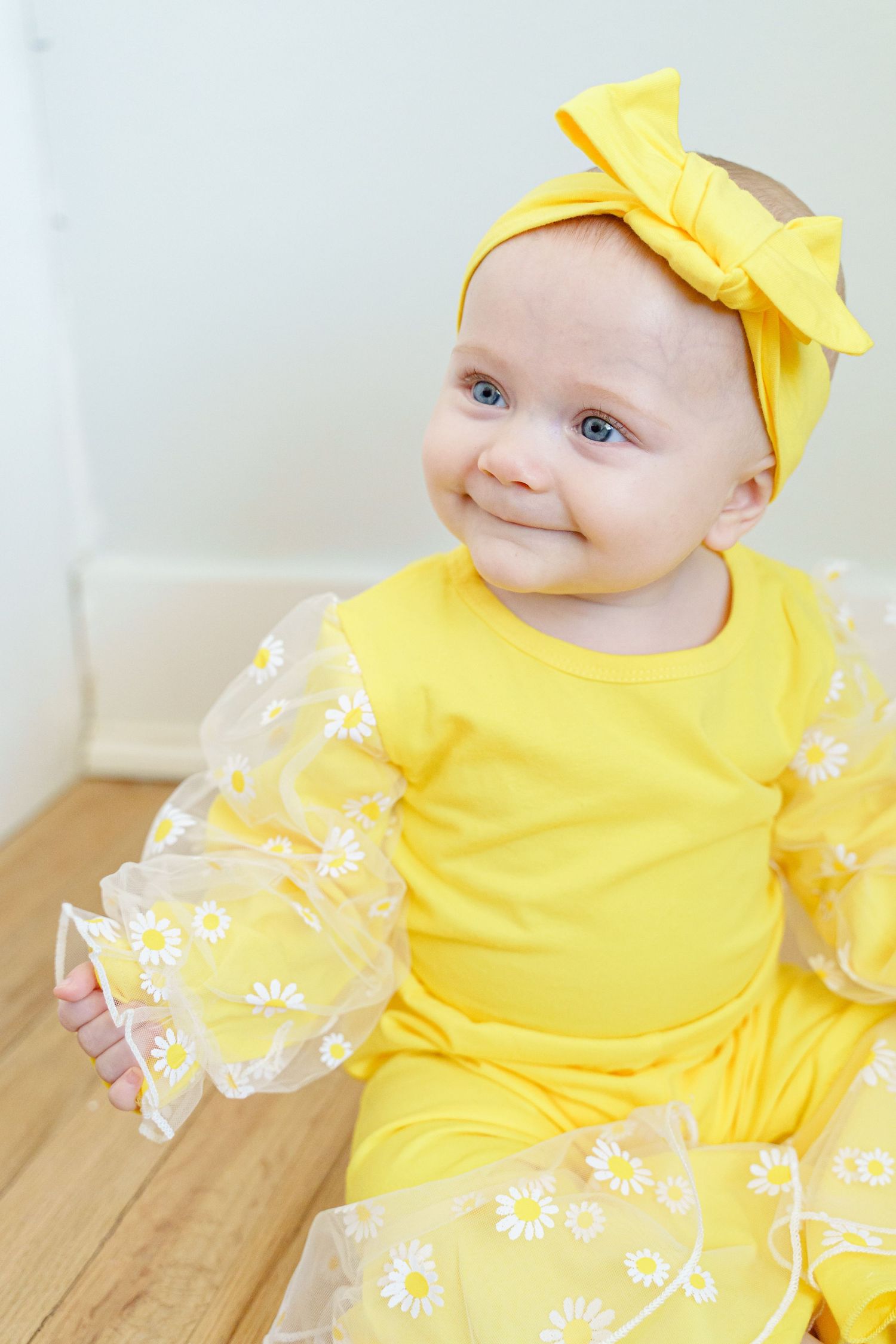 Baby wearing a bright yellow dress with daisy details and matching yellow bow headband smiles sweetly.