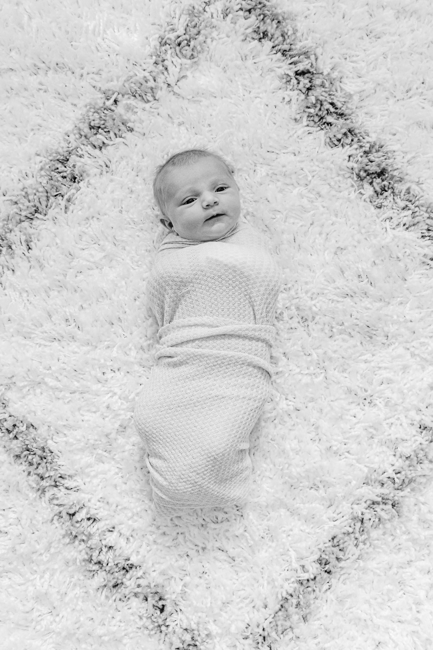 Black and white portrait of an infant swaddled in white fabric against a textured background.