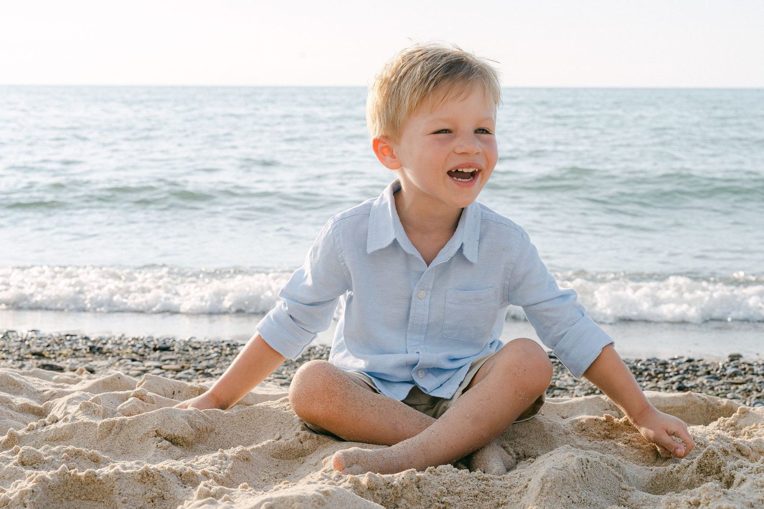 Young child playing and sitting in the sand at the beach with waves visible in the background.