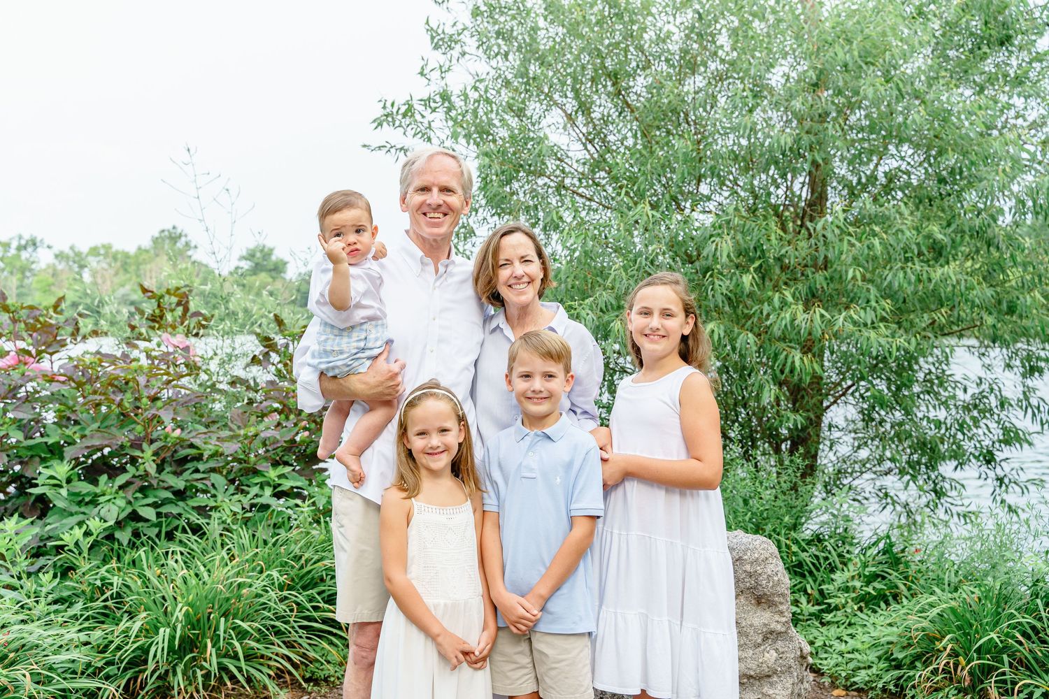 A family wearing coordinated white and blue outfits poses together outdoors in a lush green setting.