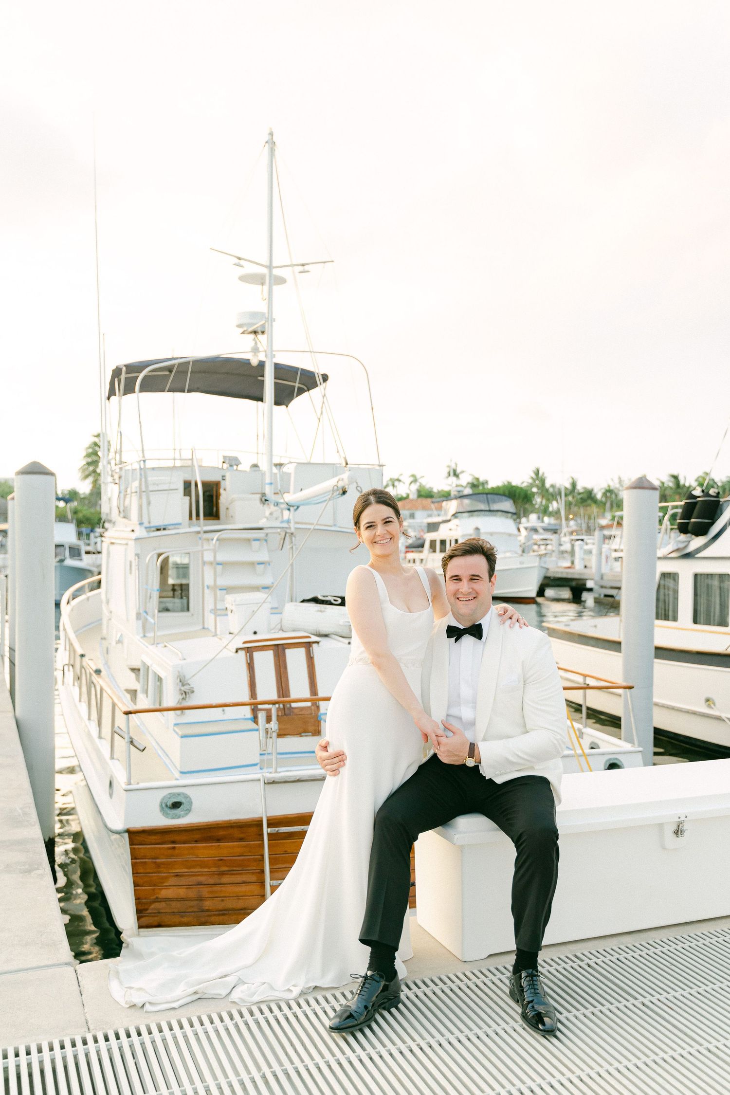 Bride and groom posing on a marina dock with luxury yachts, bride in white gown, groom in white tuxedo jacket  in Miami, FL