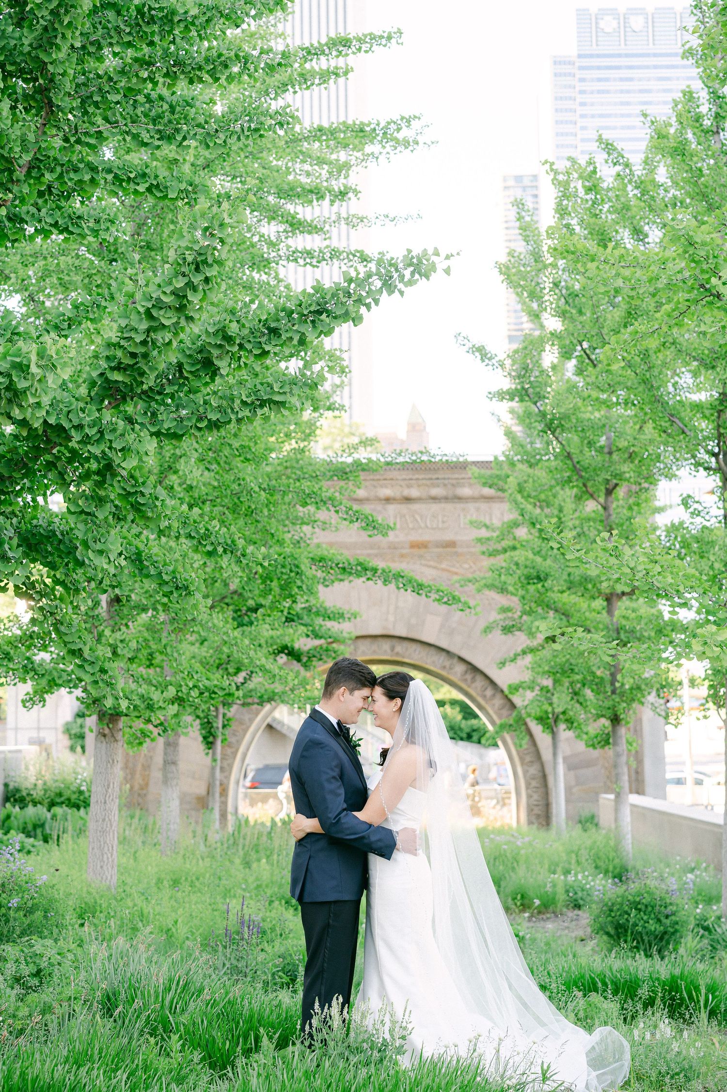 Bride and groom embrace among lush green trees with stone arches and city skyline in background in Chicago, IL