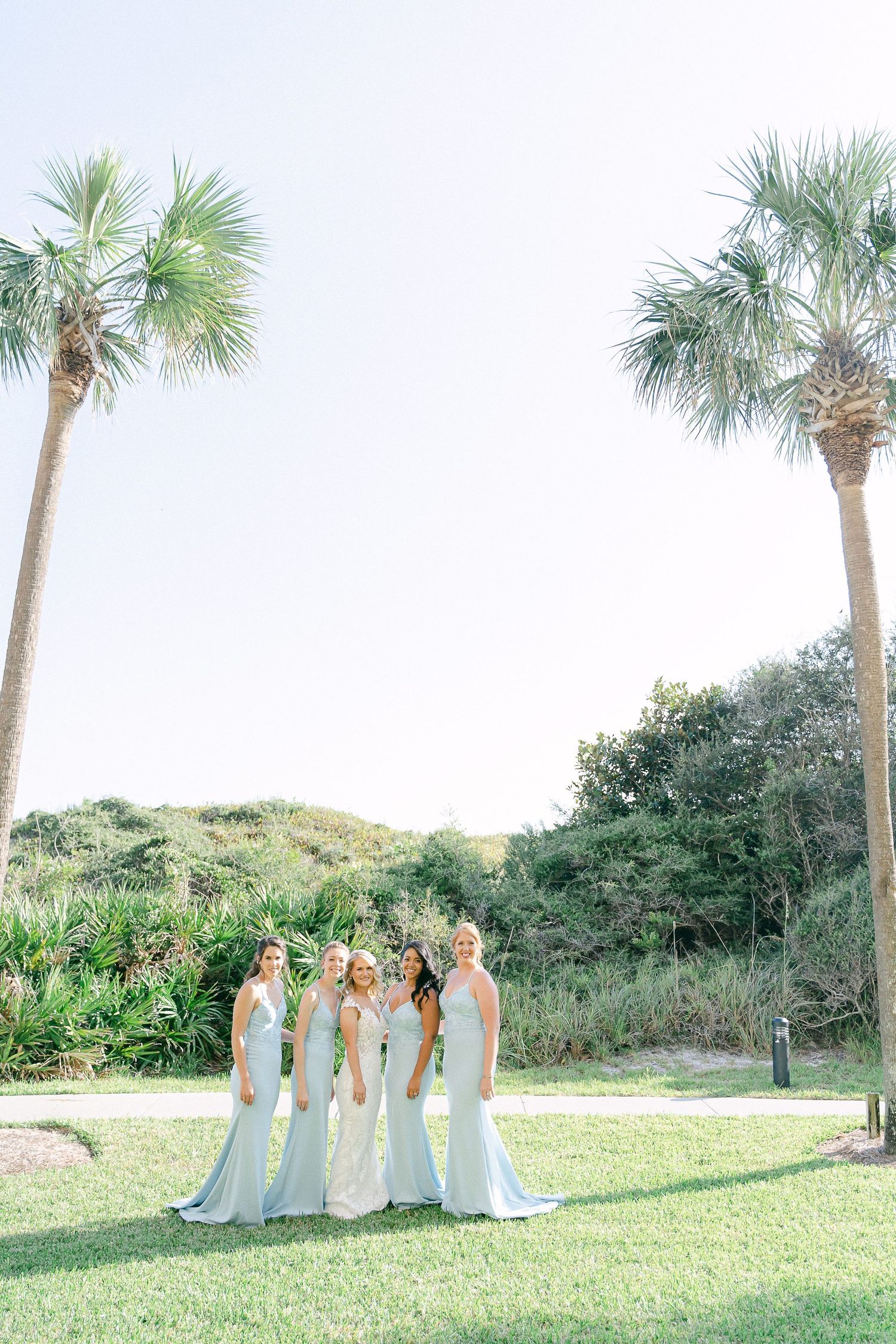 Bride and bridesmaids in light blue gowns posing on green lawn between tall palm trees at outdoor wedding venue in Destin, FL