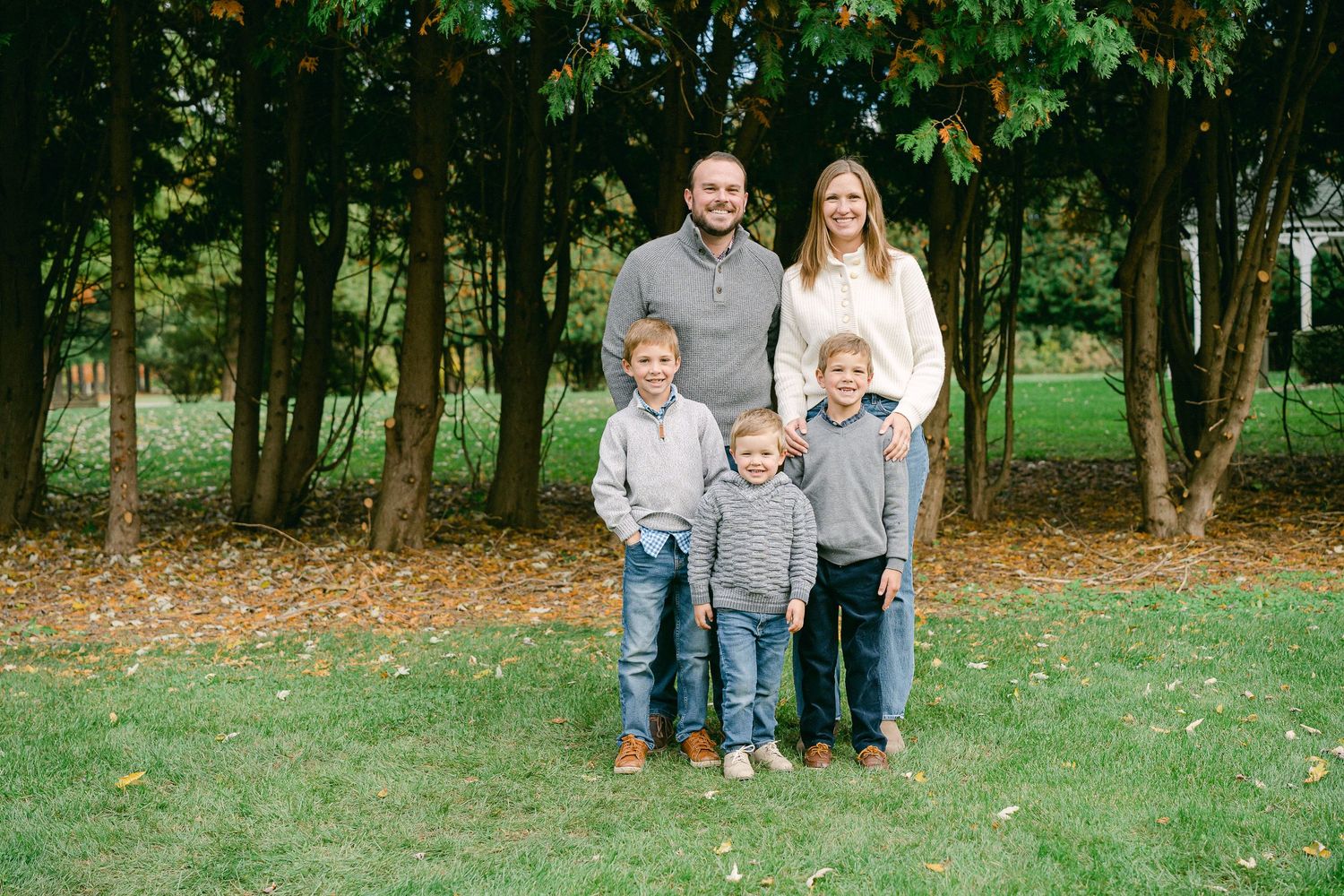 A family stands together on green grass surrounded by autumn trees during an outdoor photo session.