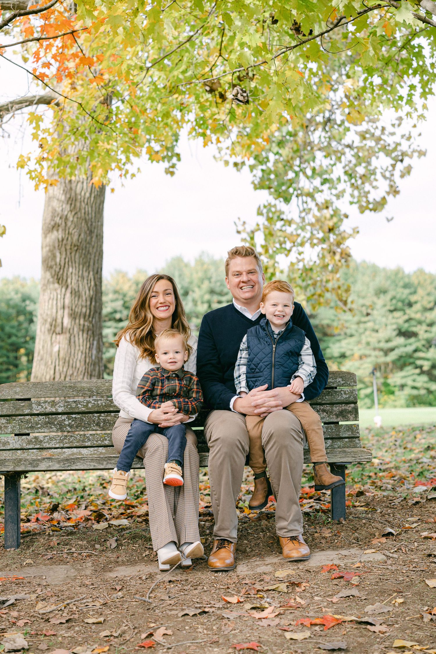 Family sits together on wooden bench surrounded by autumn leaves and trees in outdoor park setting.