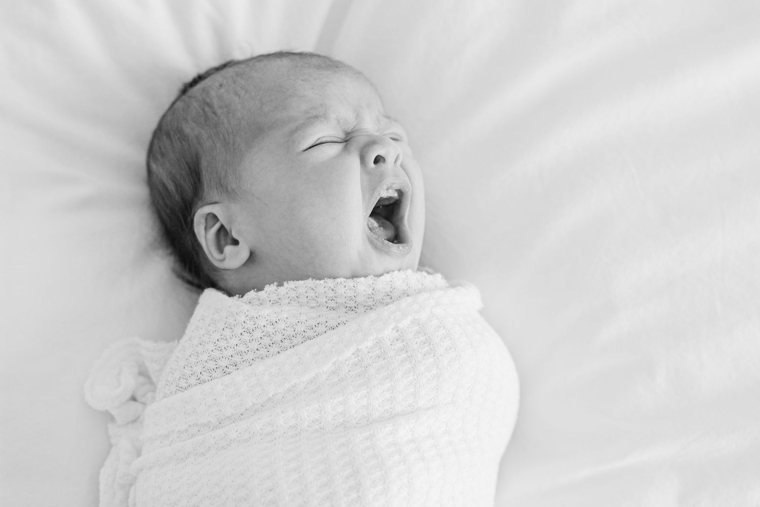 A sleeping newborn baby wrapped in white swaddle blanket lies peacefully on white bedding.