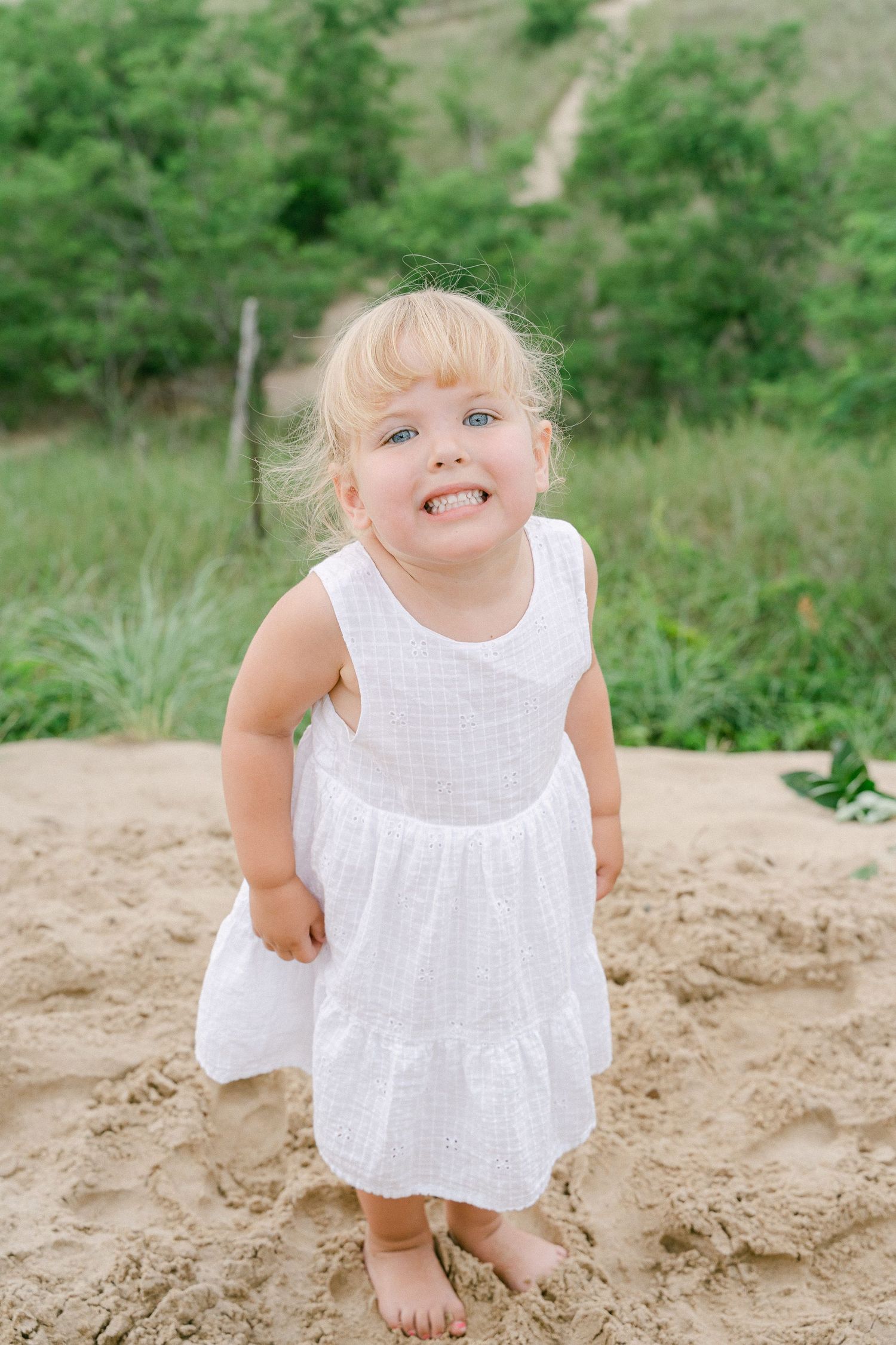 Baby wearing white eyelet dress plays in sandy area with green foliage in background.