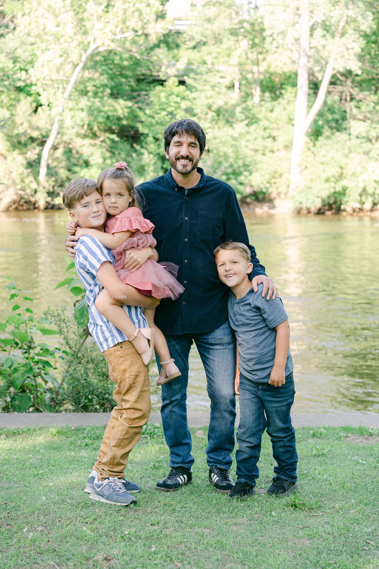 A family group poses together on grassy riverbank during an outdoor photo session.