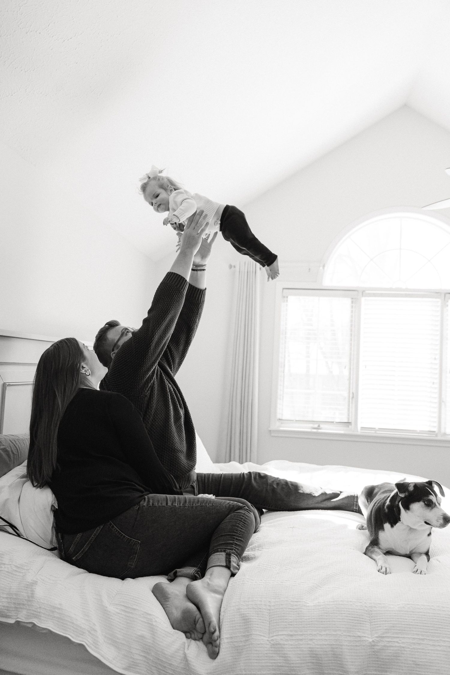 A tender black and white moment of playtime on a bed with a dog resting nearby.