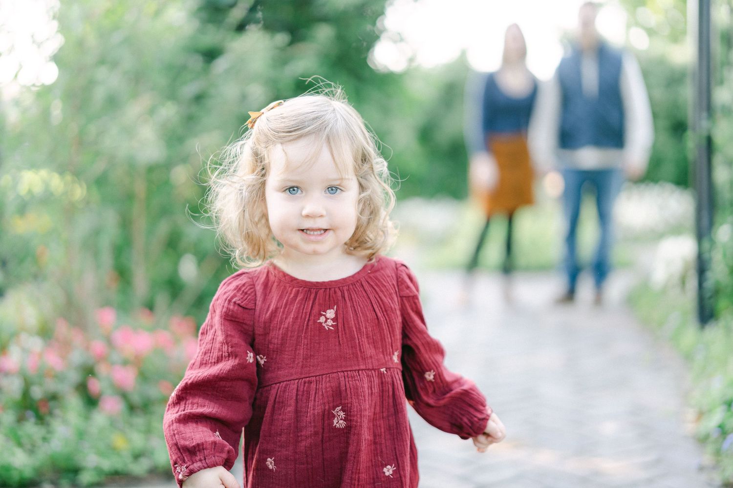 A young child wearing a burgundy dress walks along an outdoor path surrounded by greenery and flowers.