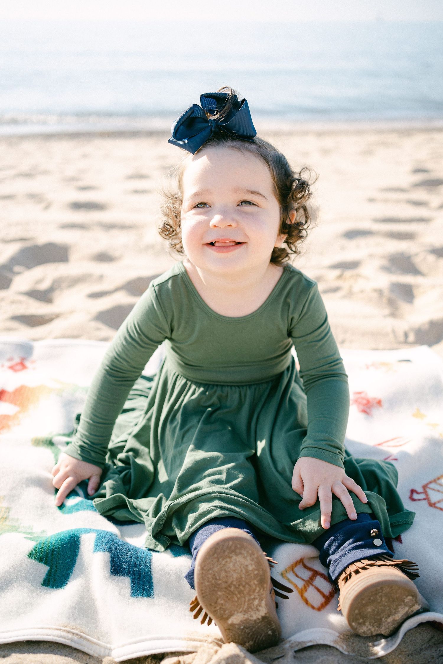 Baby in olive green dress plays on sandy beach wearing brown boots.
