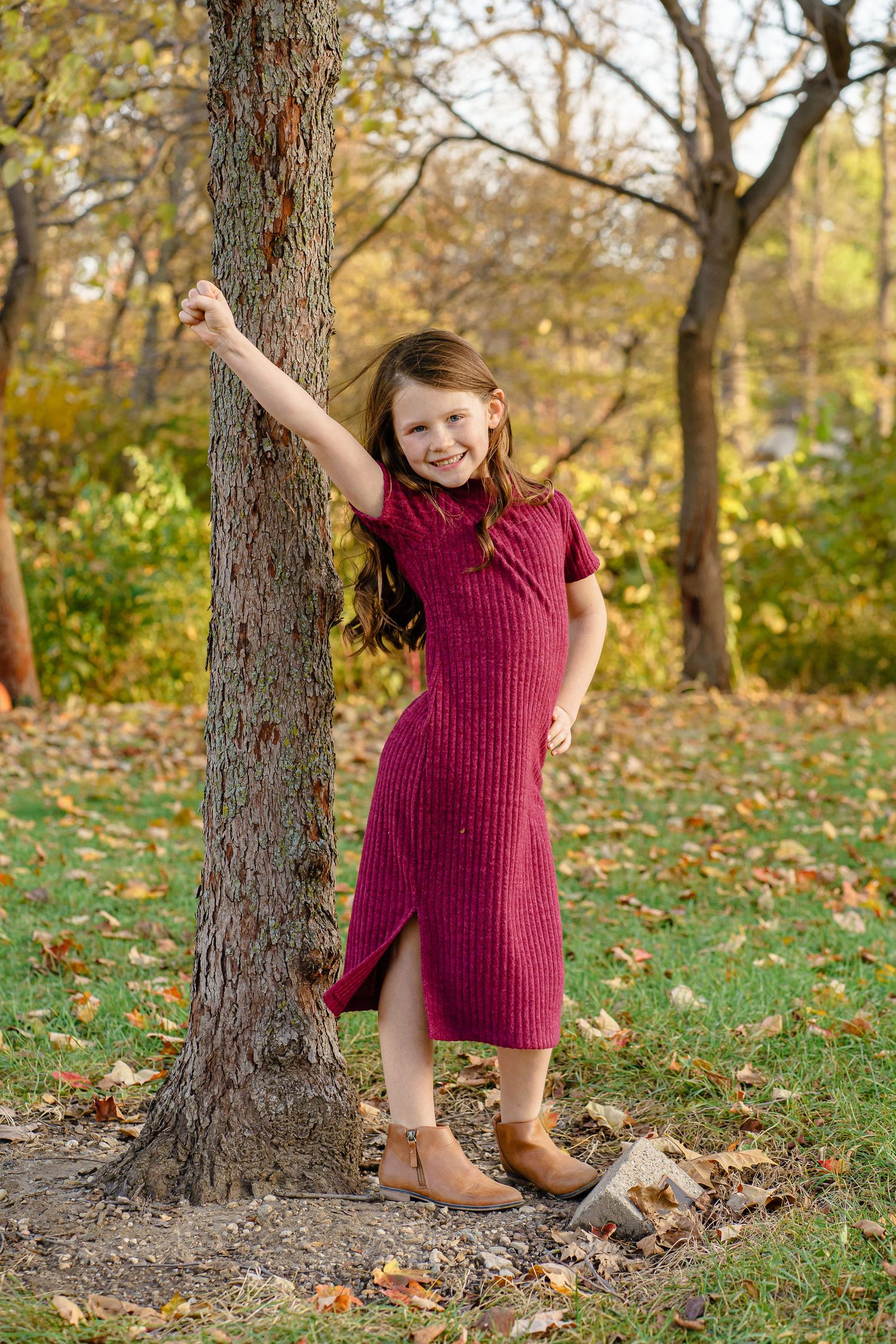 Child in burgundy sweater dress plays and poses by a tree trunk in autumn setting with fallen leaves on the ground.