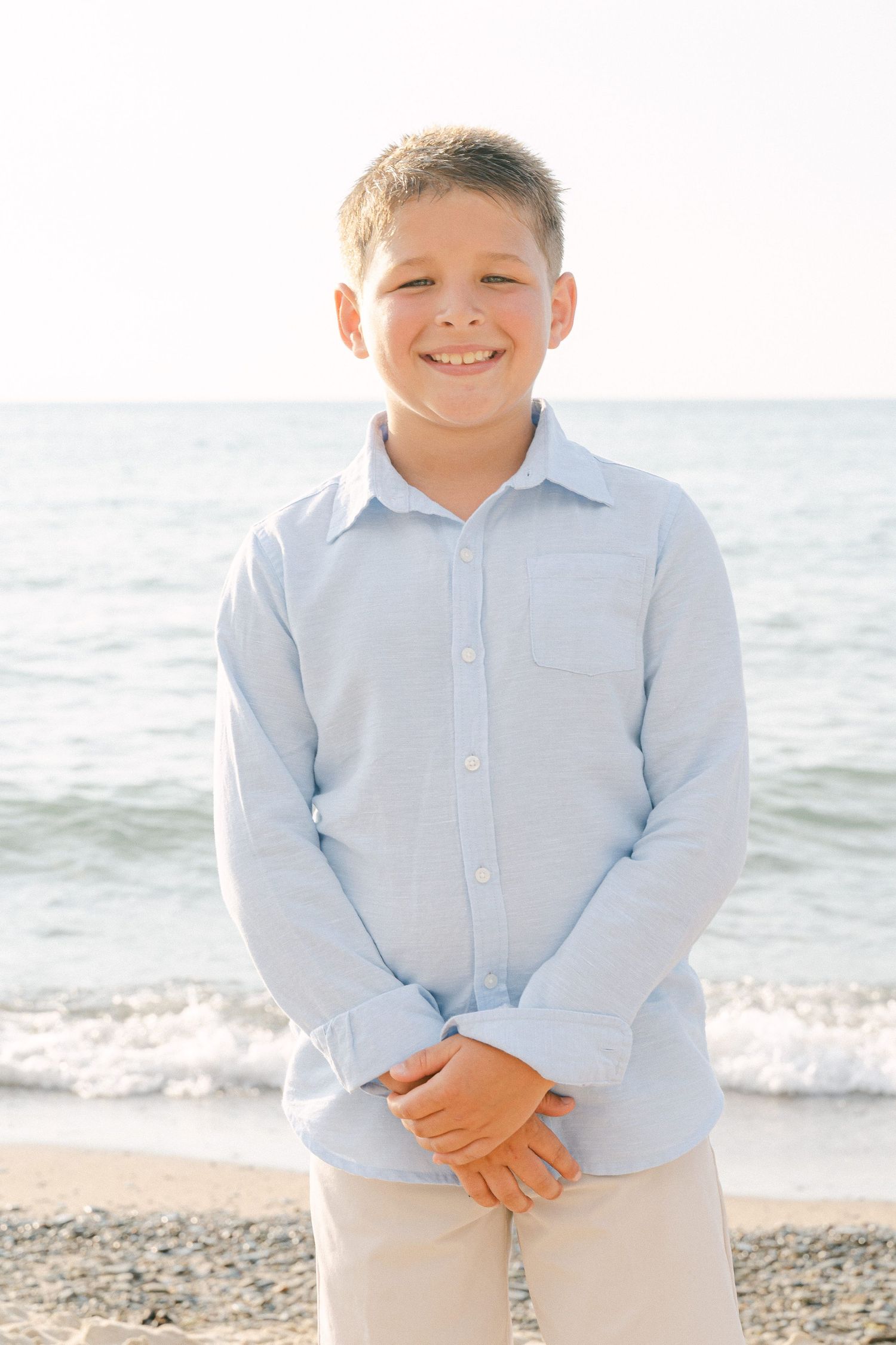 Young boy in light blue button down shirt and khaki pants standing on beach.