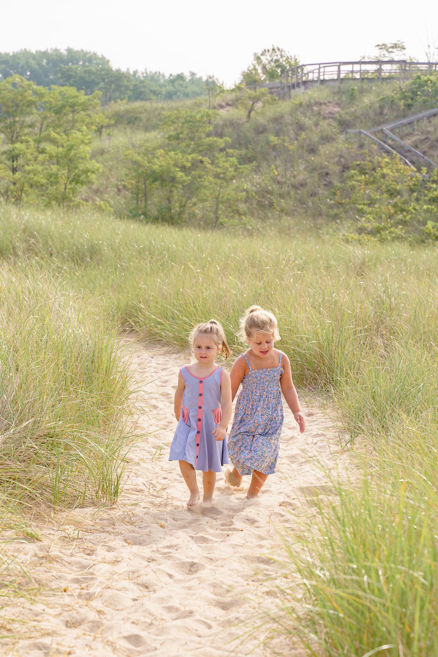 Two young children in summer dresses walking together on a sandy beach path through tall grass.