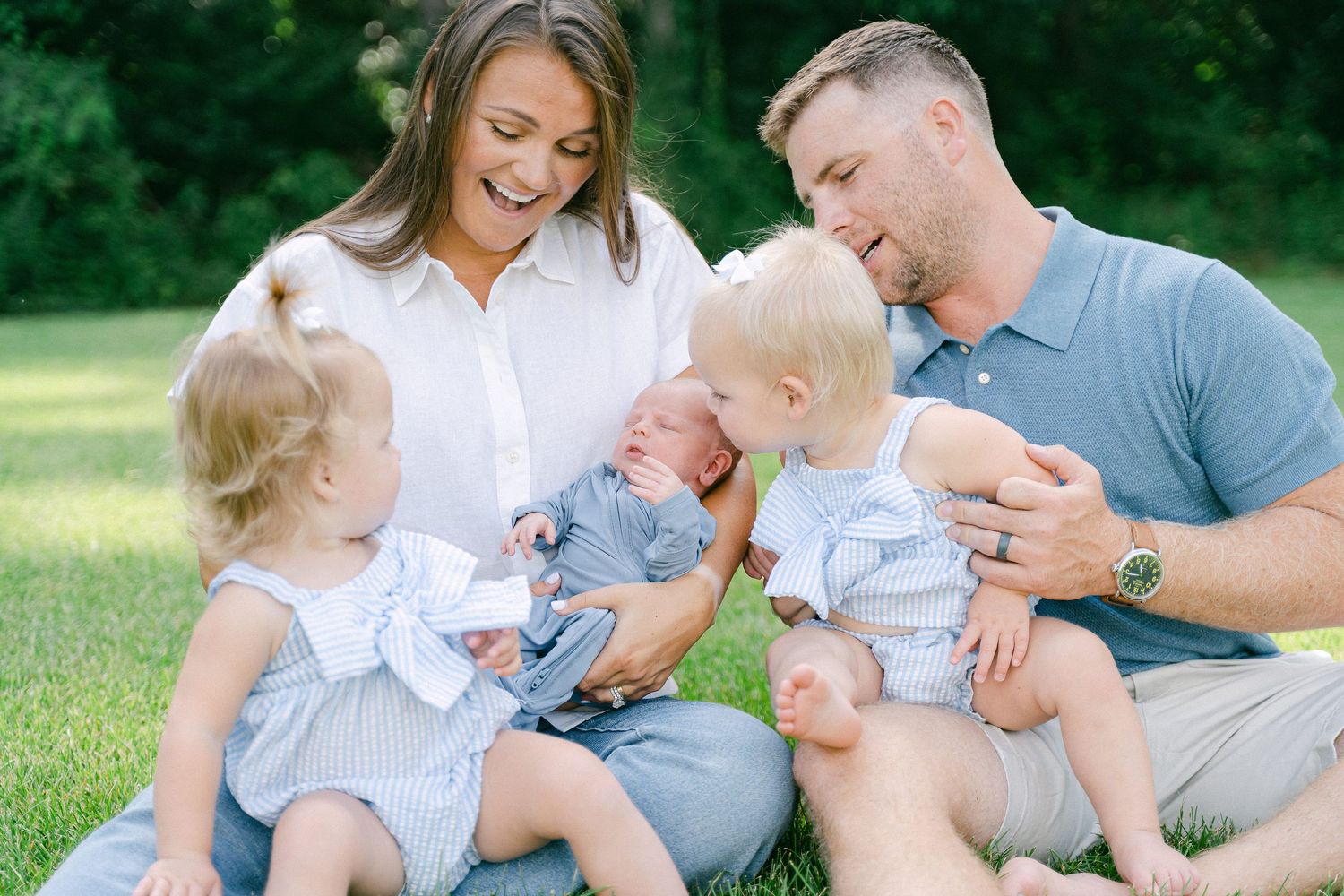 Family wearing coordinated white and gray outfits sits together on grass during outdoor photo session.