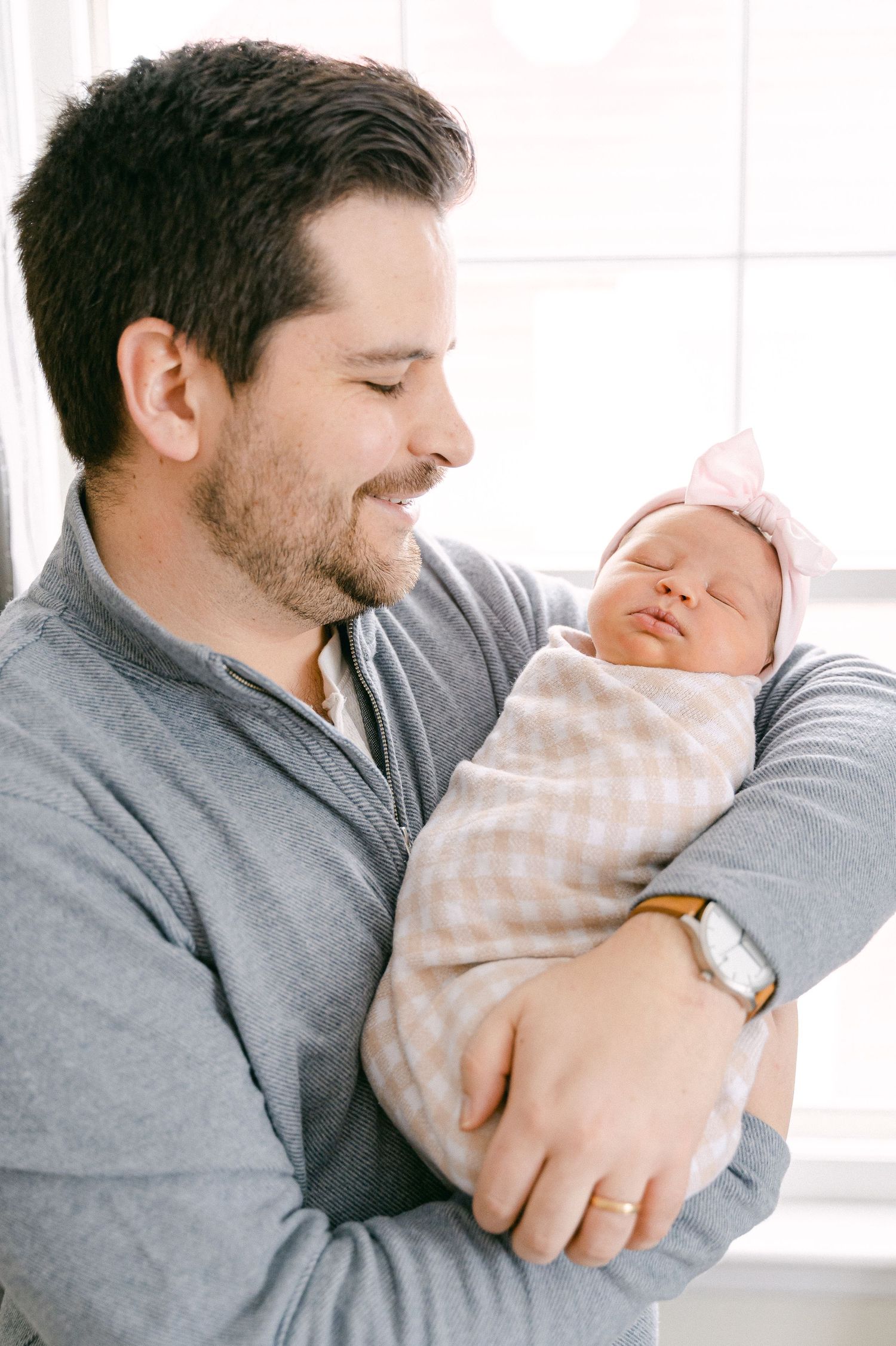 A tender moment between a parent in a gray sweater holding and gazing lovingly at their sleeping newborn wrapped in stripes.