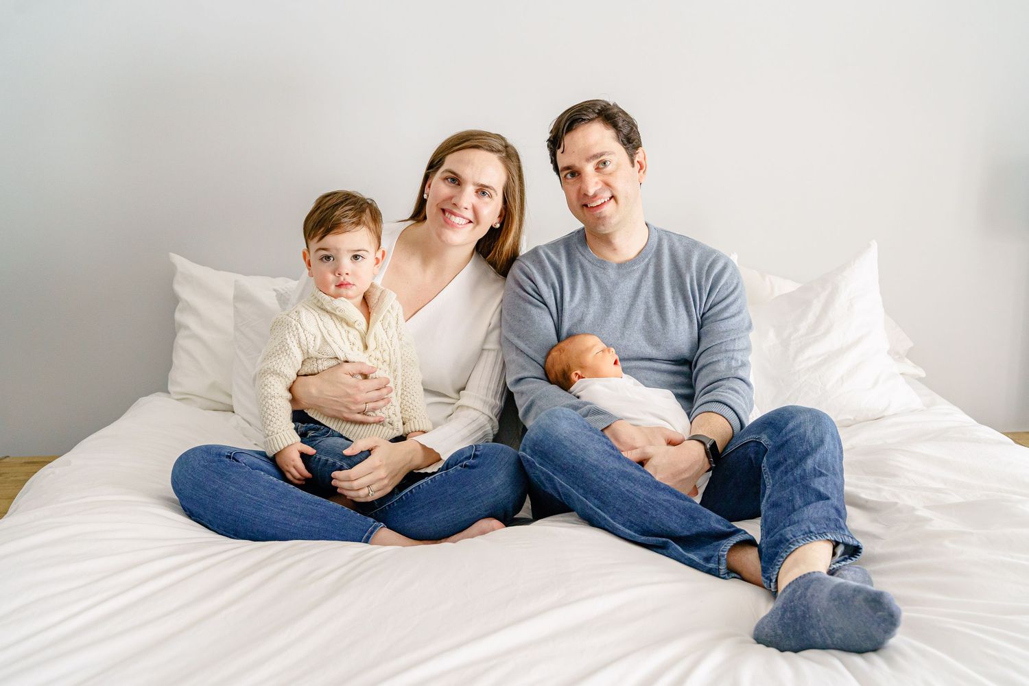 A family relaxes together on a white bed wearing casual clothes in a bright minimalist setting.