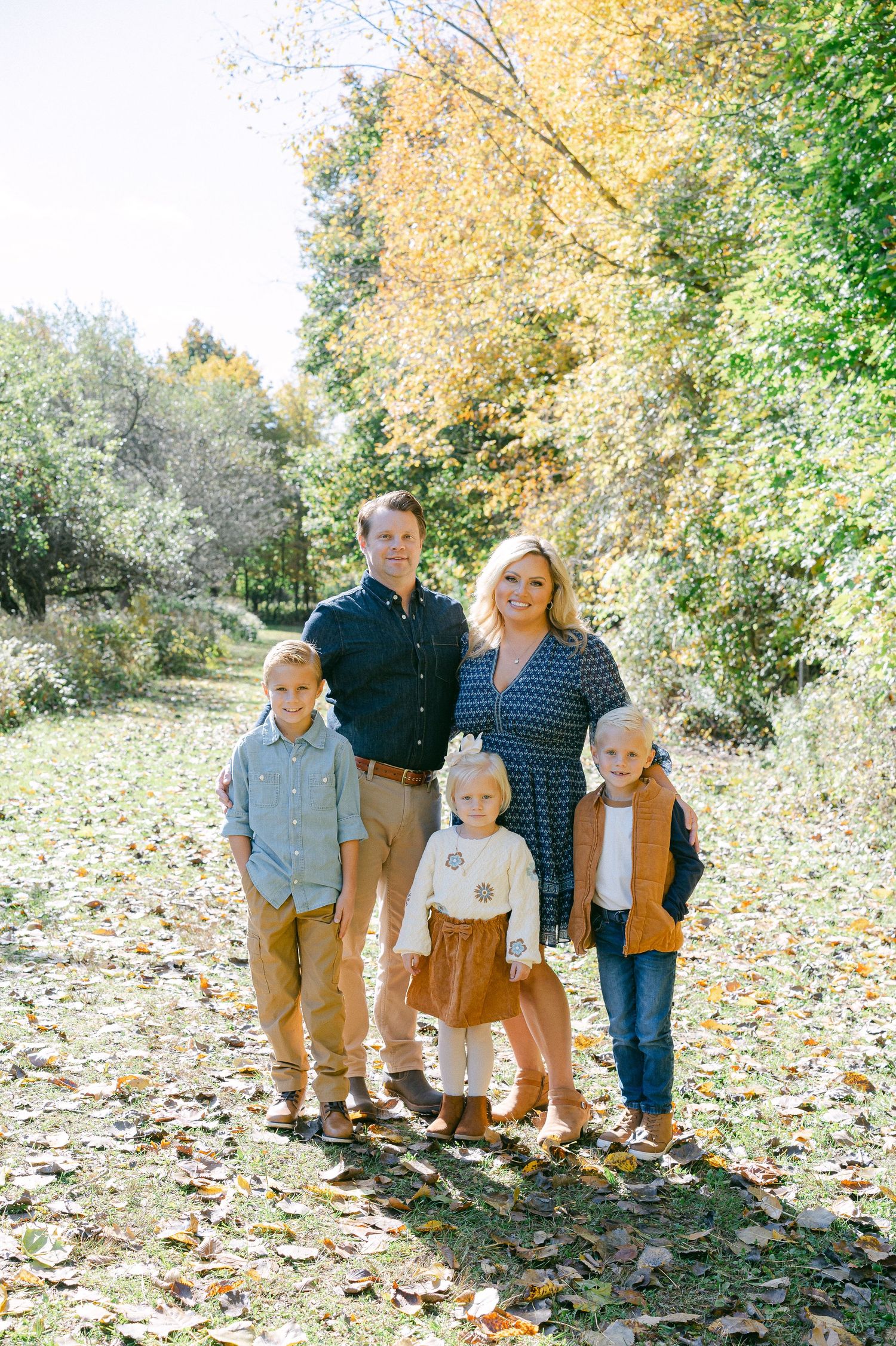 A family group poses outdoors on a path covered in colorful fall leaves with trees in the background.