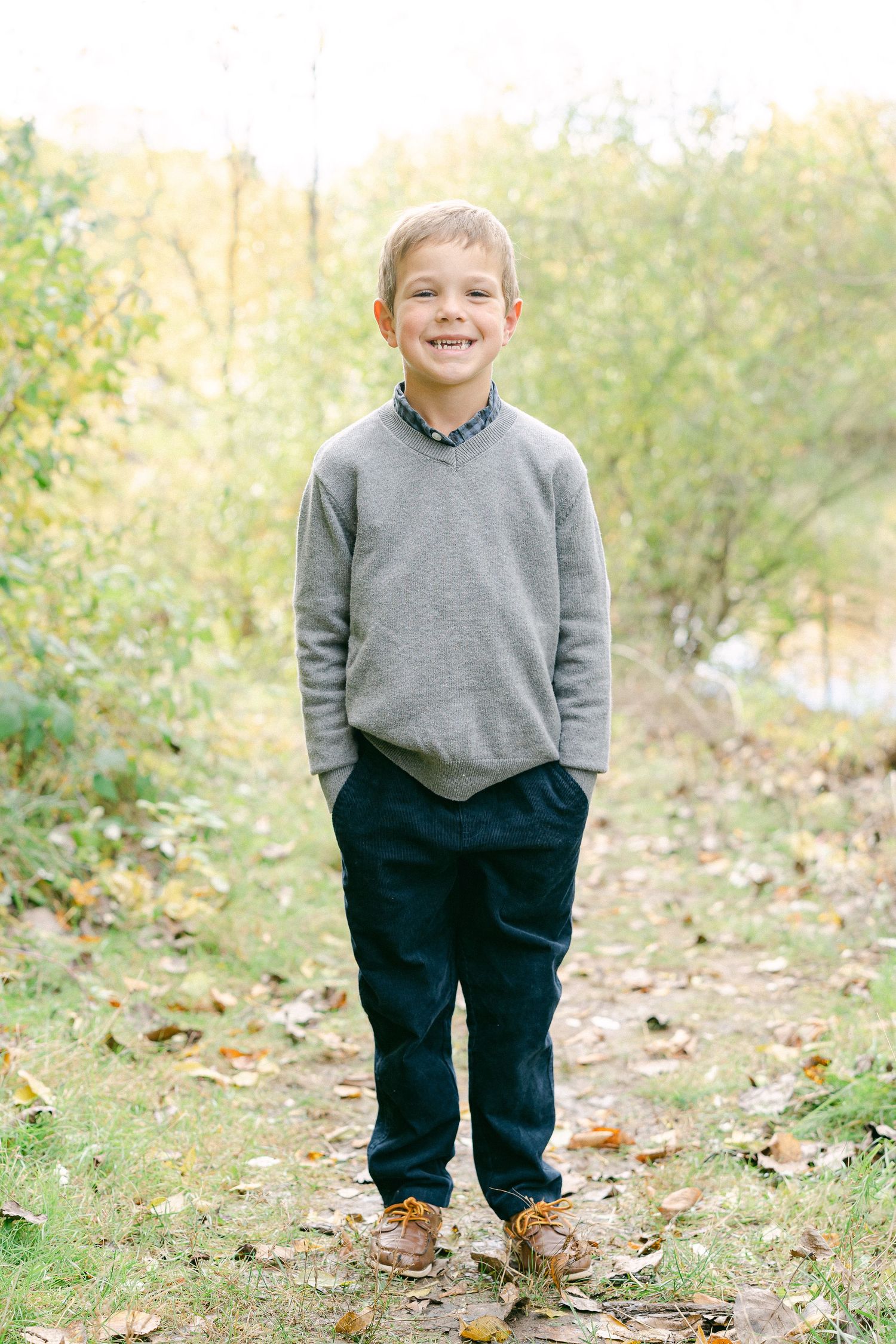 A person in a grey sweater and dark pants stands on fallen leaves during an autumn outdoor photo session.