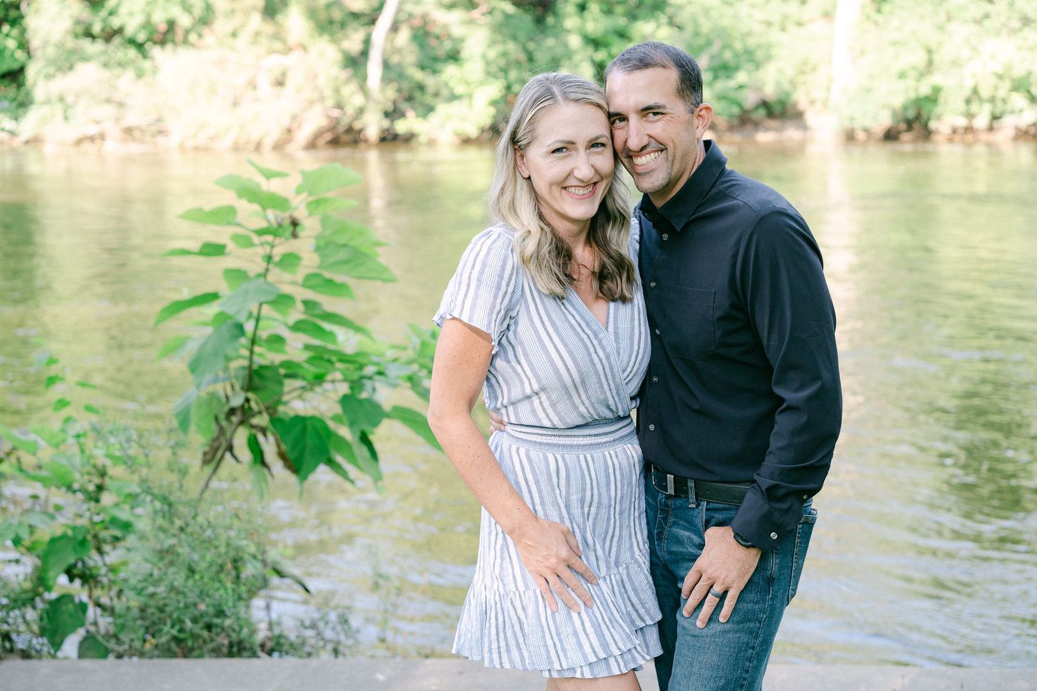 A couple poses together by a tranquil river bank on a sunny day, with green foliage in the foreground.