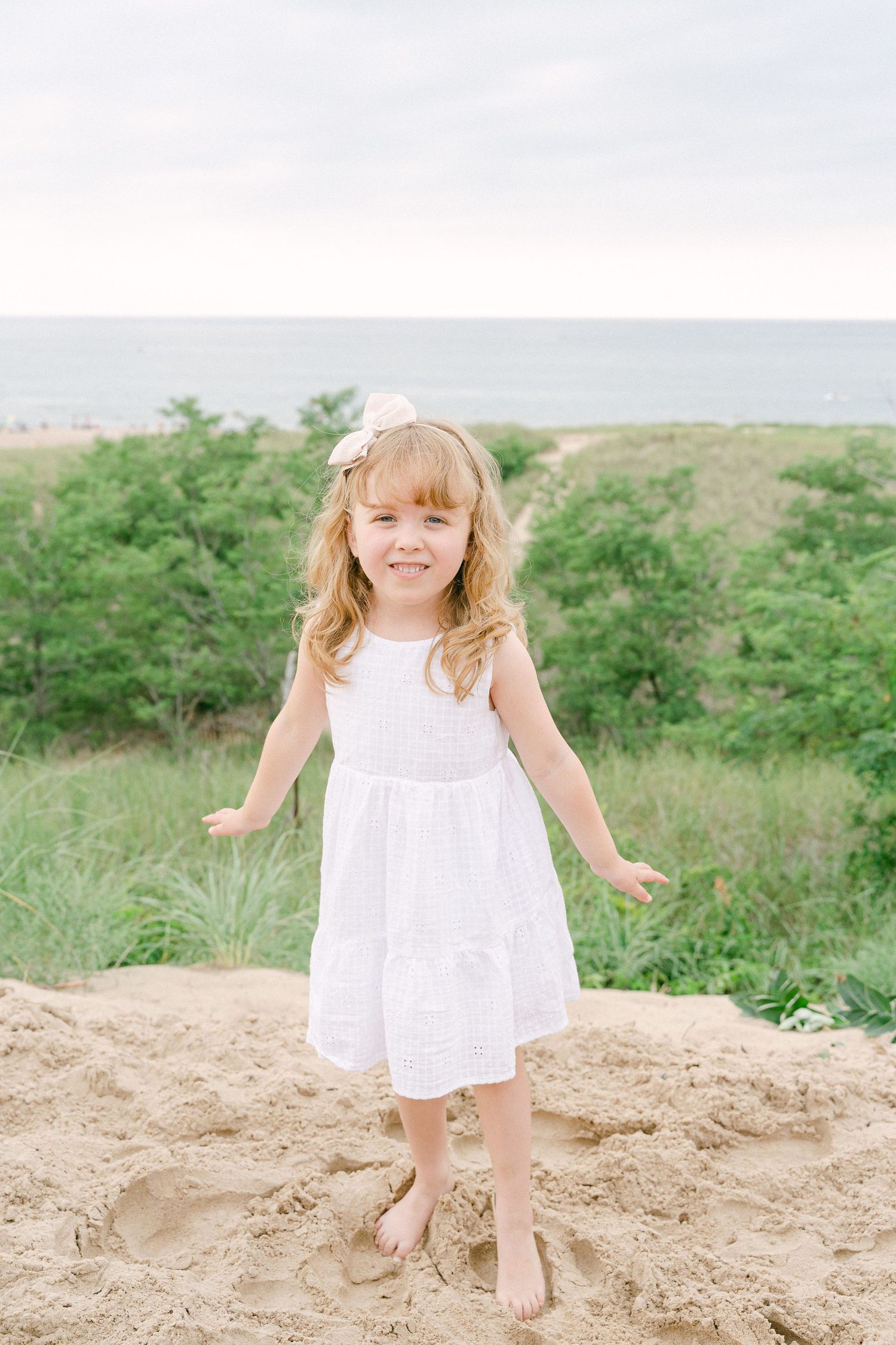 A child in a white summer dress plays on a sandy hill in a natural outdoor setting.