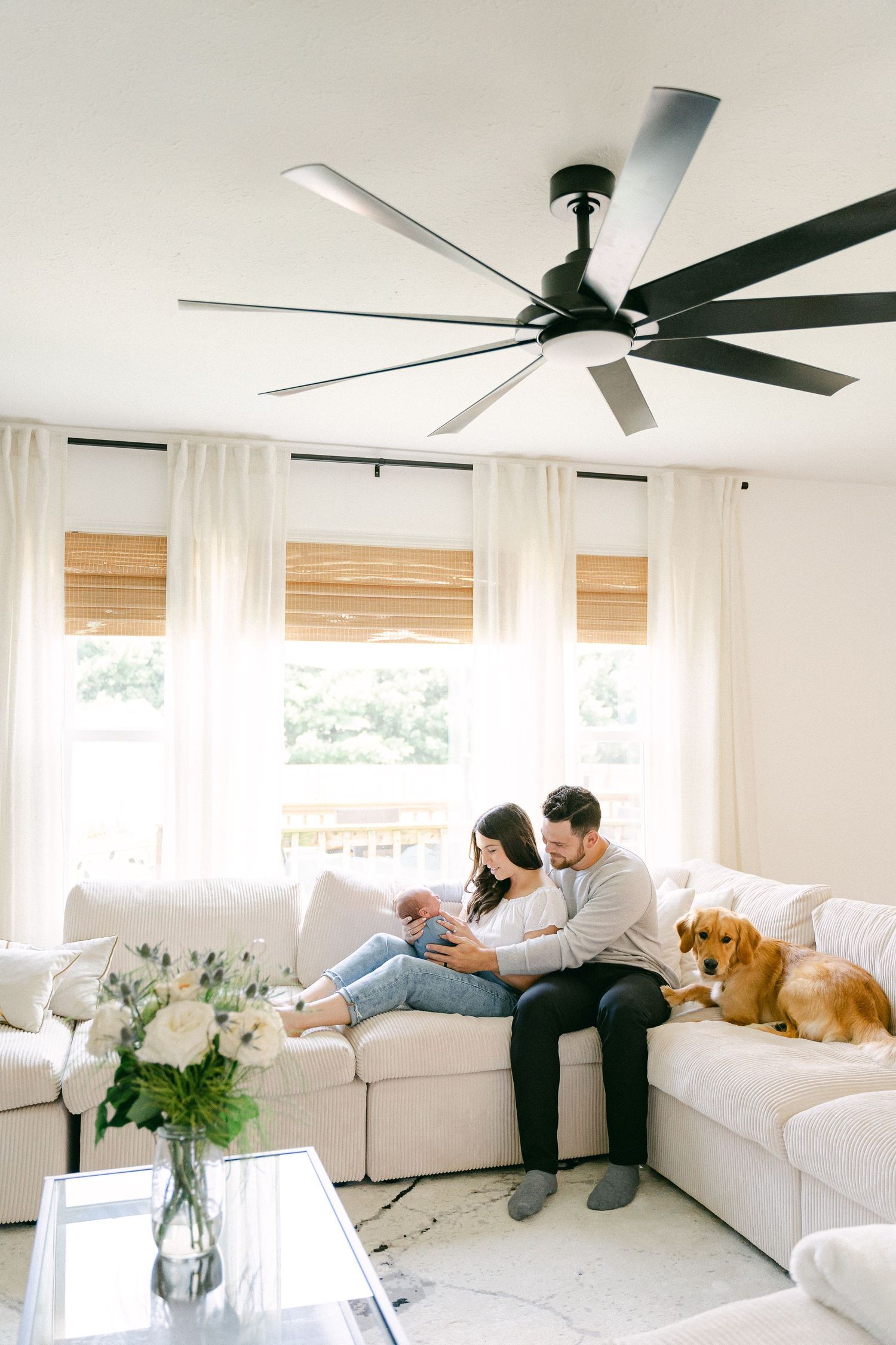 A family relaxes together on a white sectional sofa in a bright, modern living room with bamboo shades.