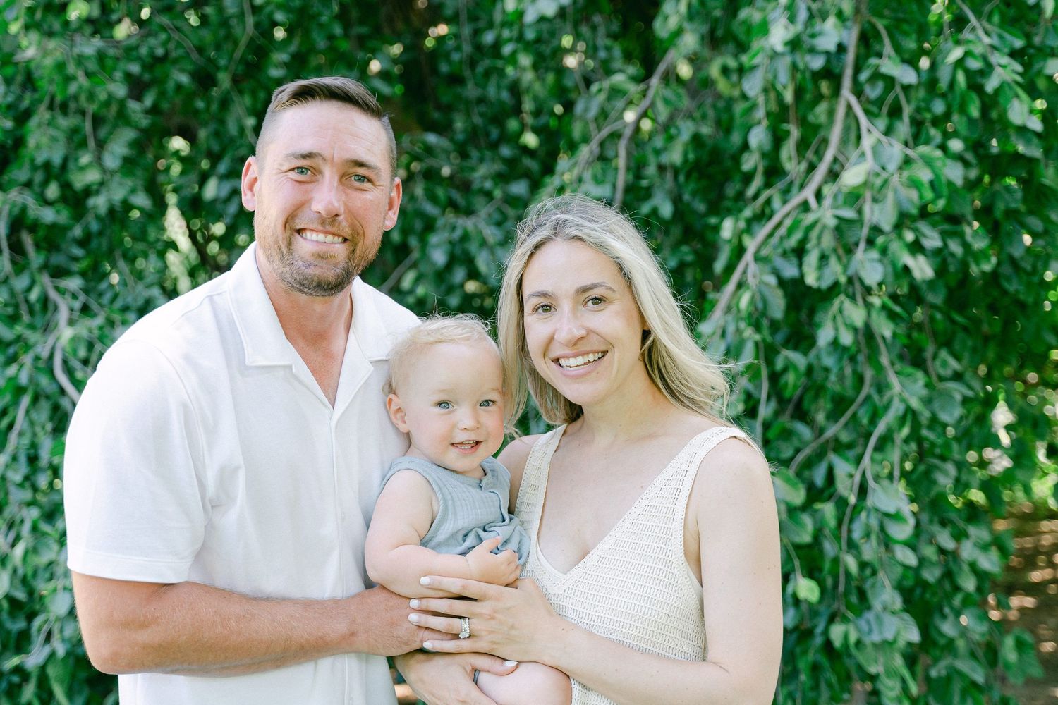 Family in white outfits poses together outdoors against lush green foliage on a sunny day.