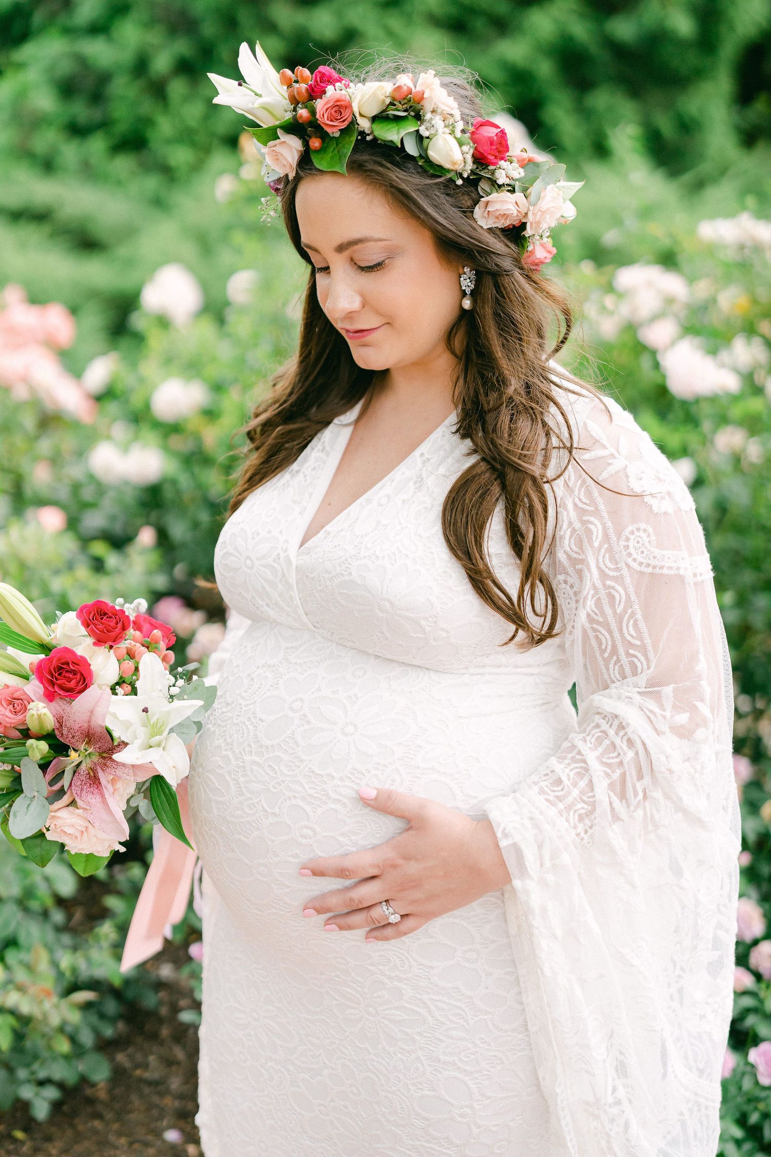 A pregnant woman in a white lace dress and floral crown poses in a garden setting with pink roses.