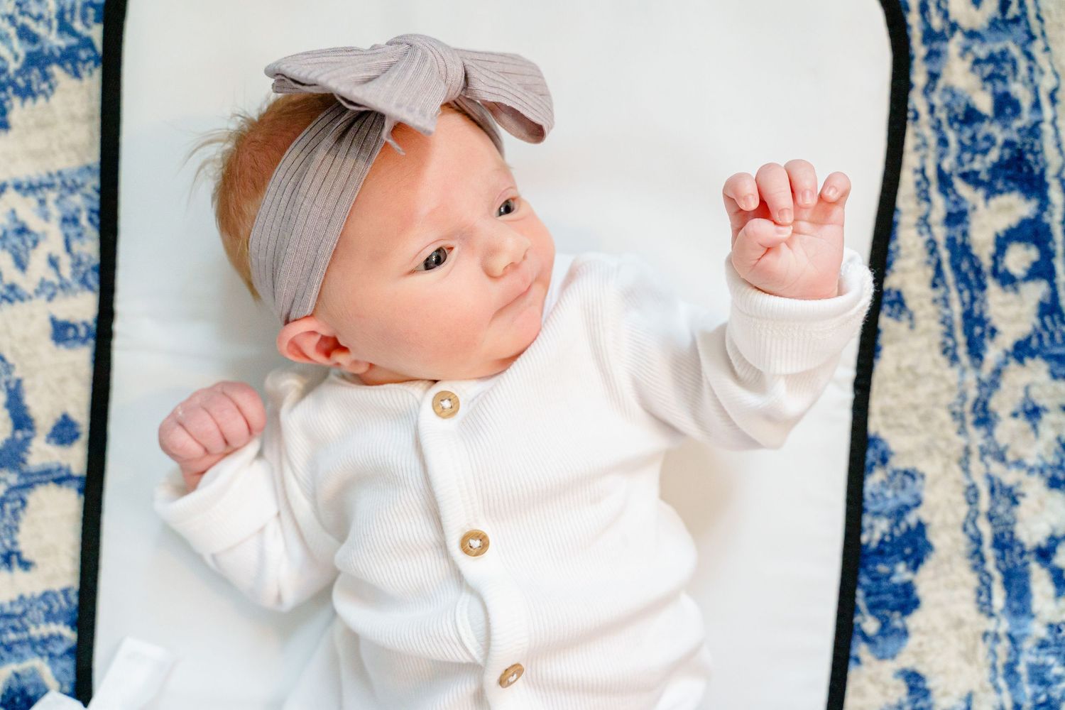 A baby wearing a white onesie and bow headband lies on a blue and white patterned background.