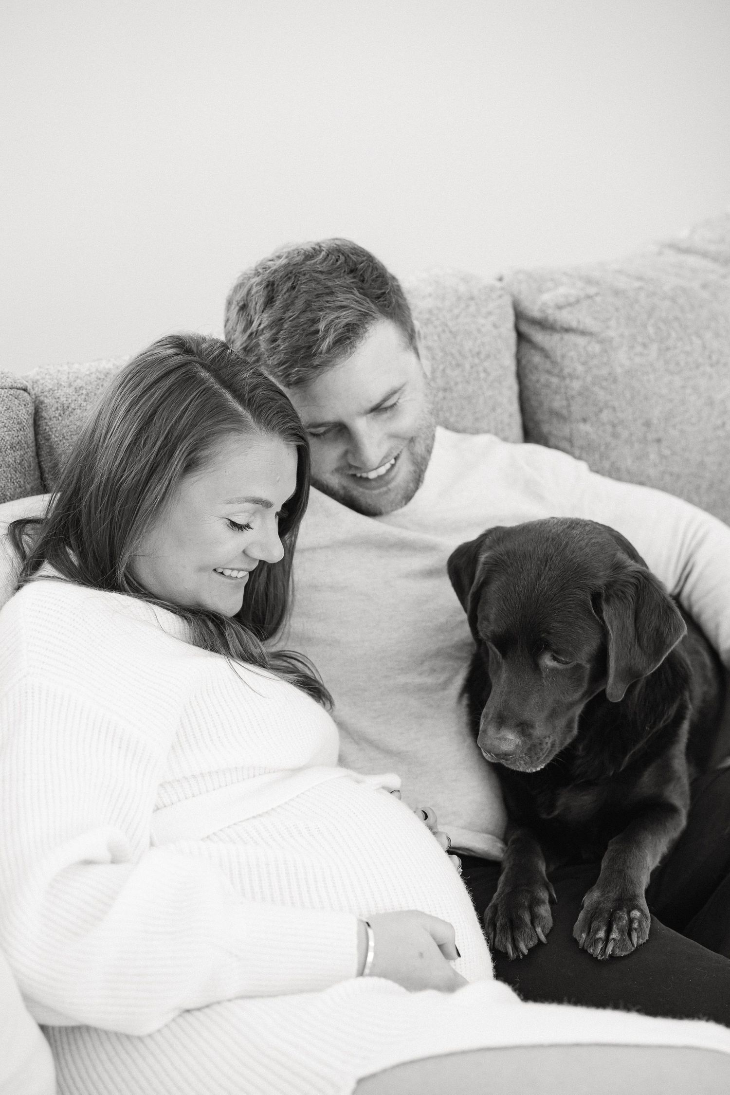 Black and white photo of couple cuddling with their chocolate lab on couch.
