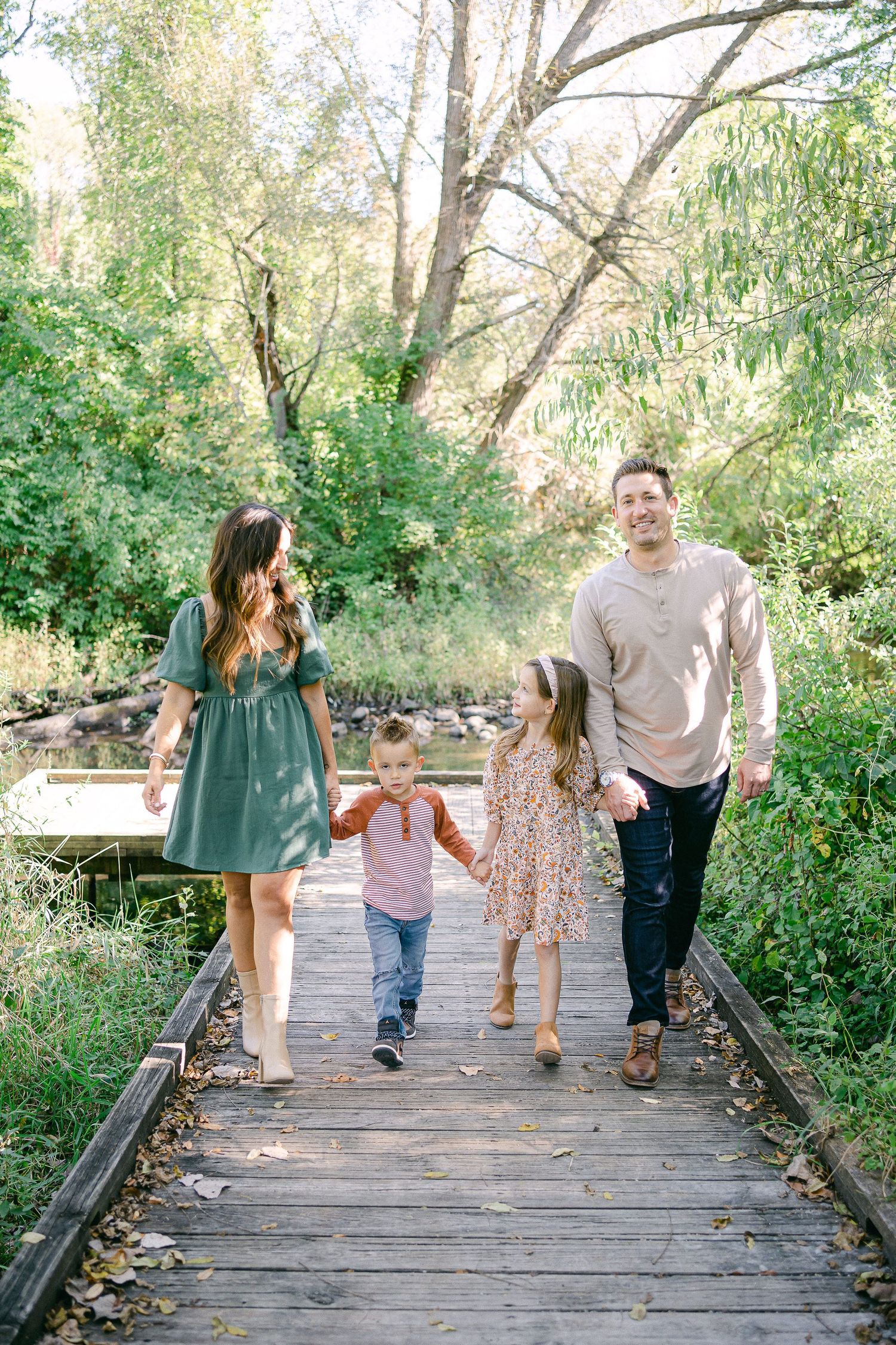 A family walks together on a wooden boardwalk path through lush green foliage on a sunny afternoon.